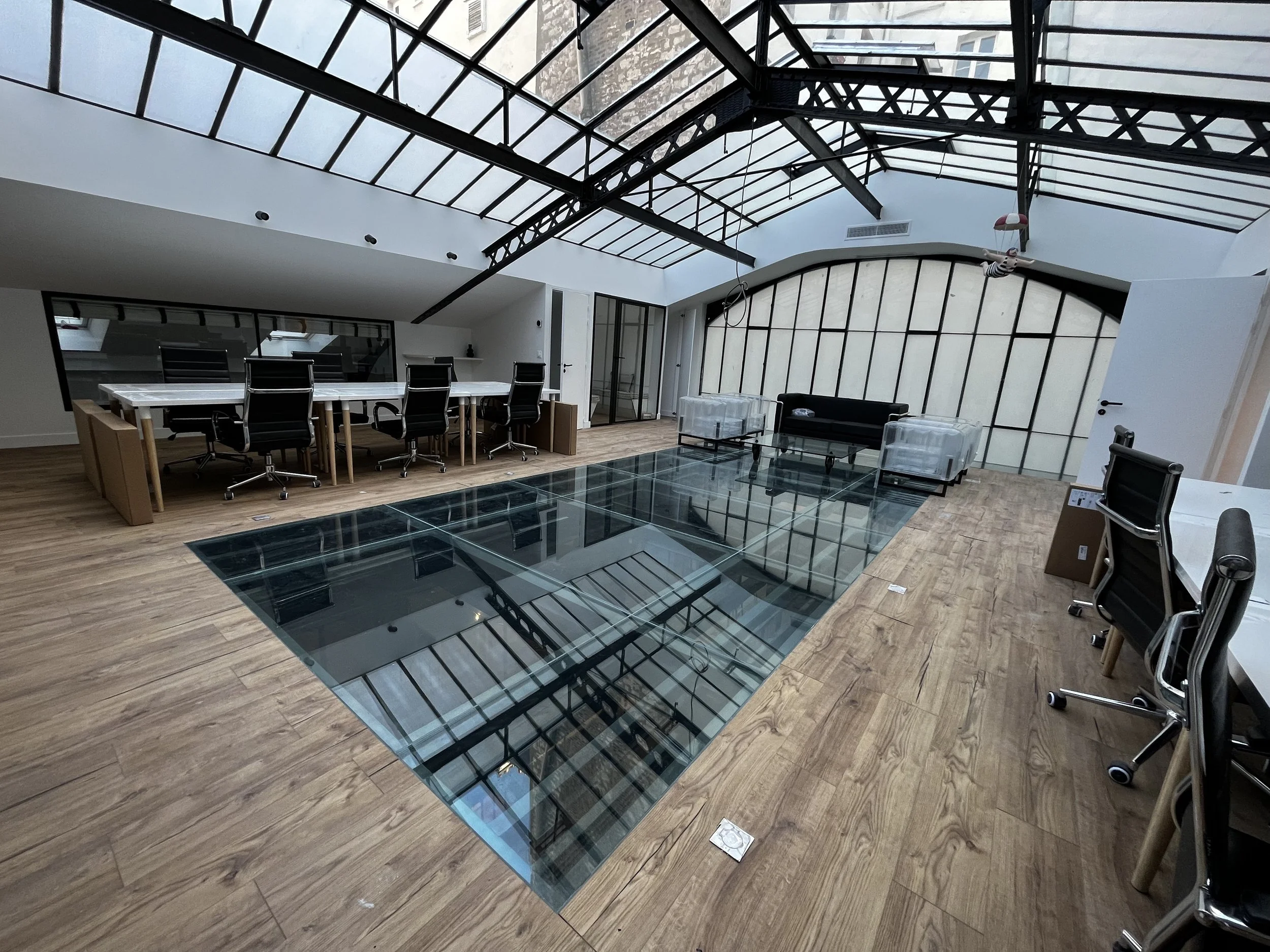 Une salle de réunion moderne avec un plafond en verre, des chaises de bureau, une table en verre, un canapé noir et des parois en verre séparant différentes zones.