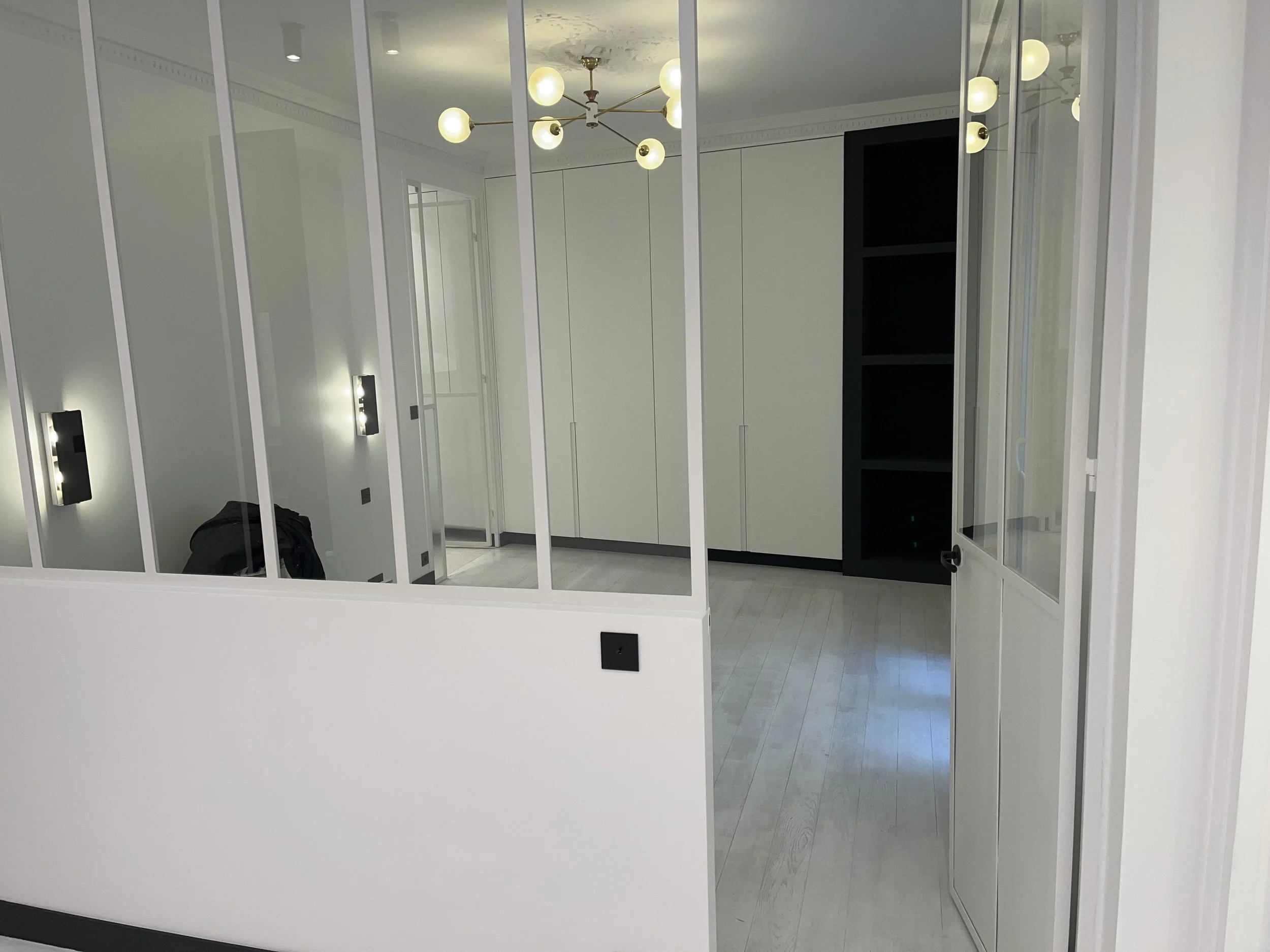 Salle moderne avec un plafond blanc décoré, un chandelier avec plusieurs boules lumineuses, des murs blancs avec des lampes murales allumées, des armoires blanches et un sol en revêtement de sol clair.
