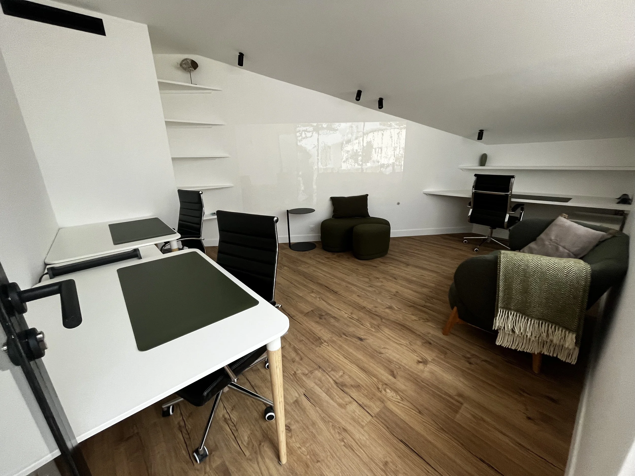 Salle de réunion moderne avec bureau en blanc, chaises noires, étagères blanches au mur et canapé vert avec coussin, sol en bois.