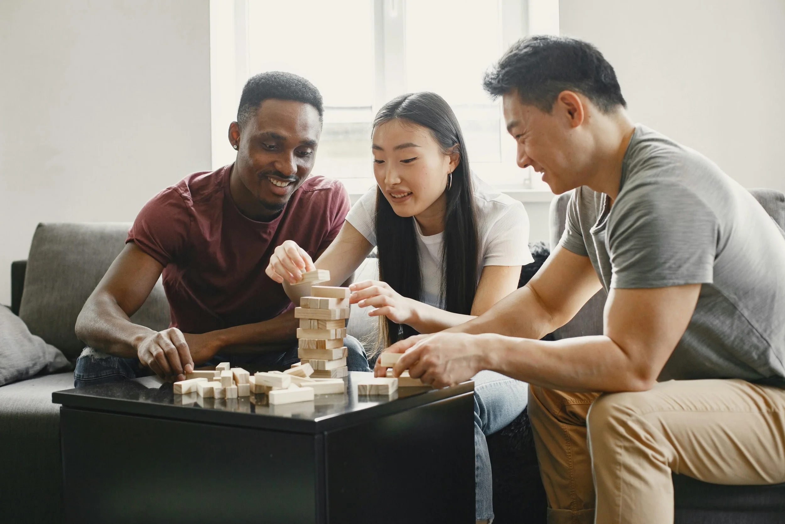 Three friends playing a wooden block game on a black table in a bright living room.
