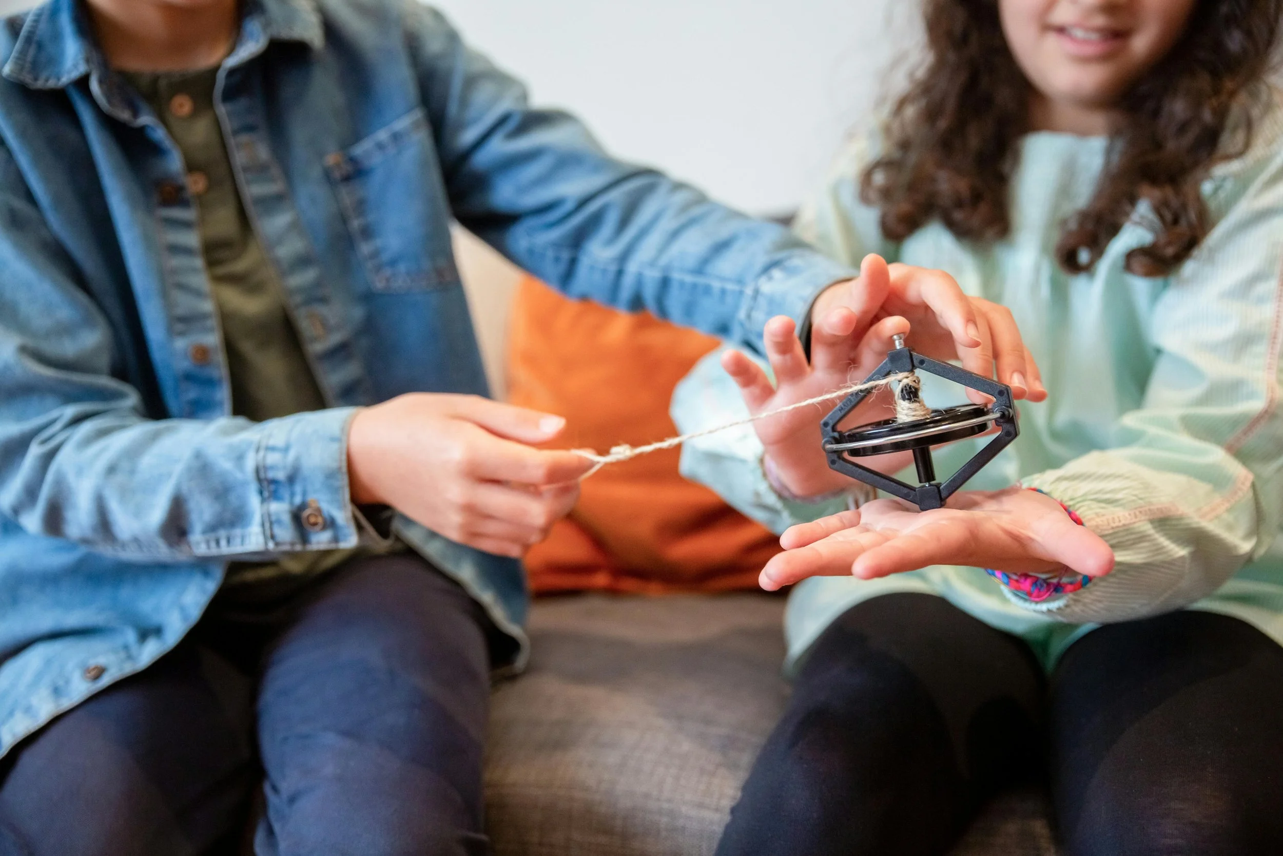 Two people are playing with a fidget spinner, which is held on one person's hand. They are sitting on a couch, and their faces are partially visible.
