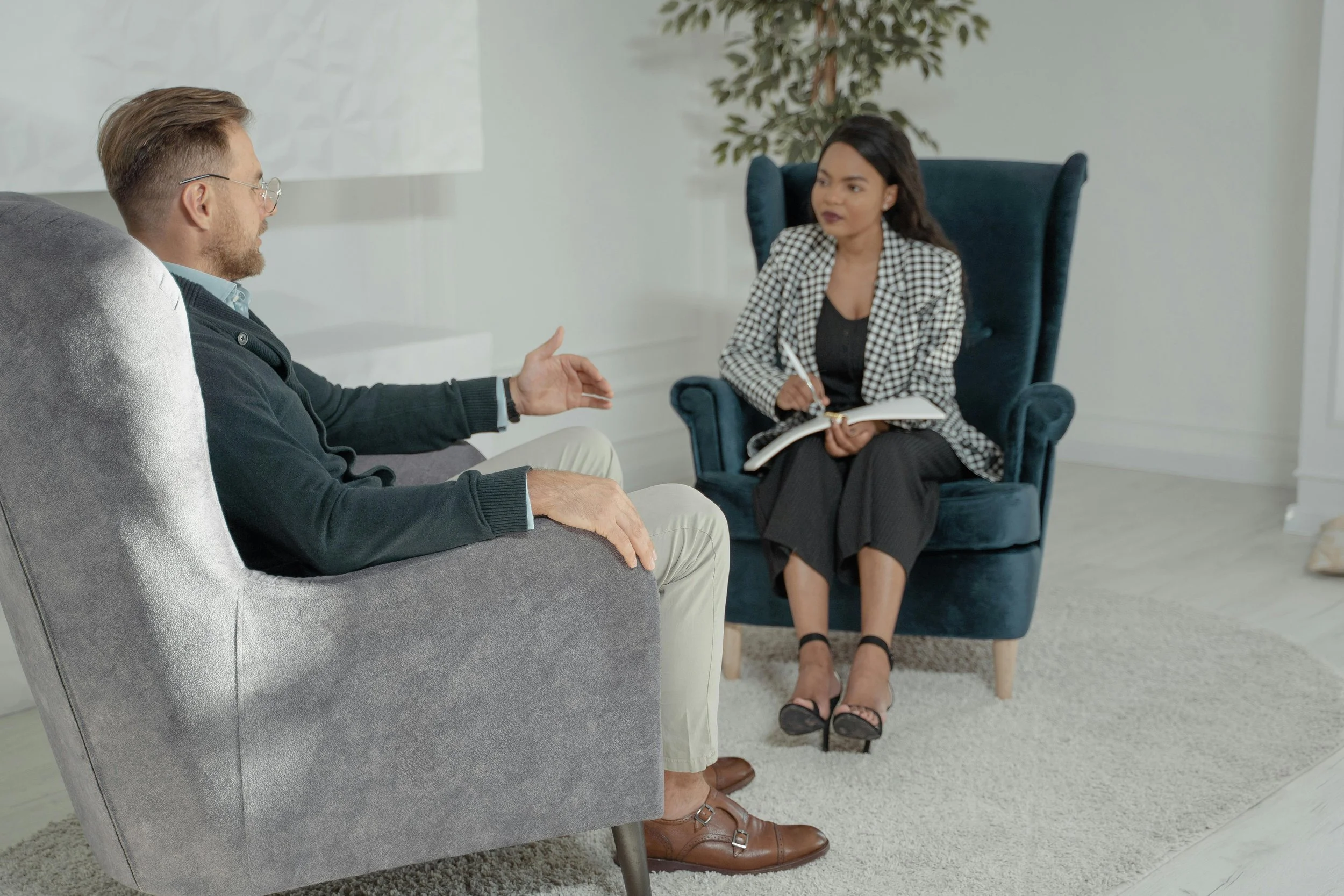A man and a woman having a conversation in a therapy session or counseling setting. The man is sitting on a gray armchair, gesturing with his hand, and the woman is sitting on a blue armchair, taking notes.