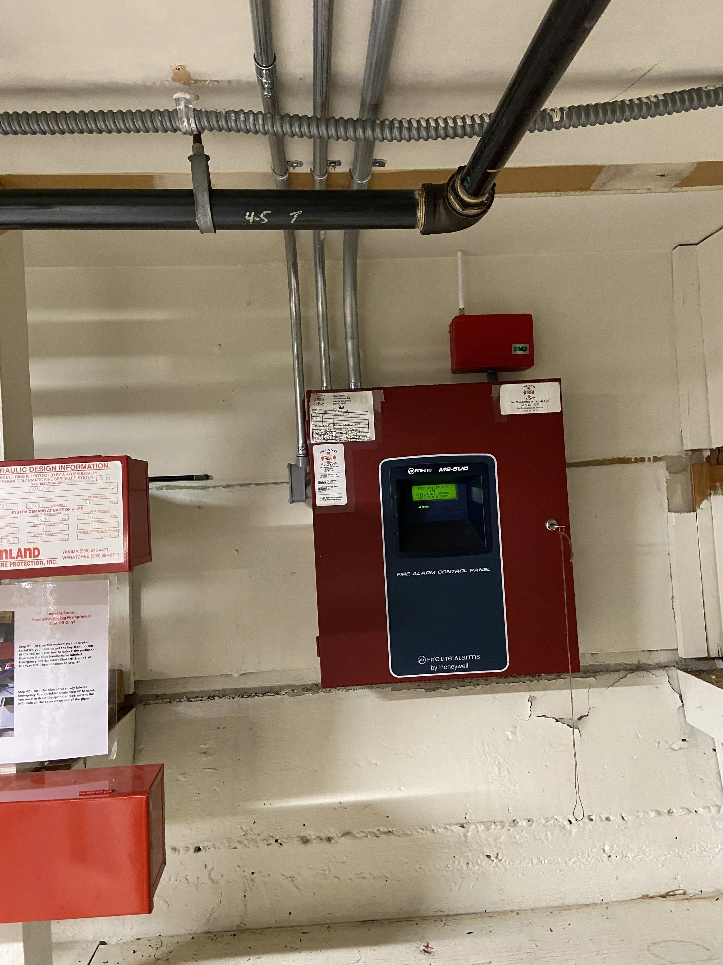 A red fire alarm control panel mounted on a white wall, with pipes and wiring above it.