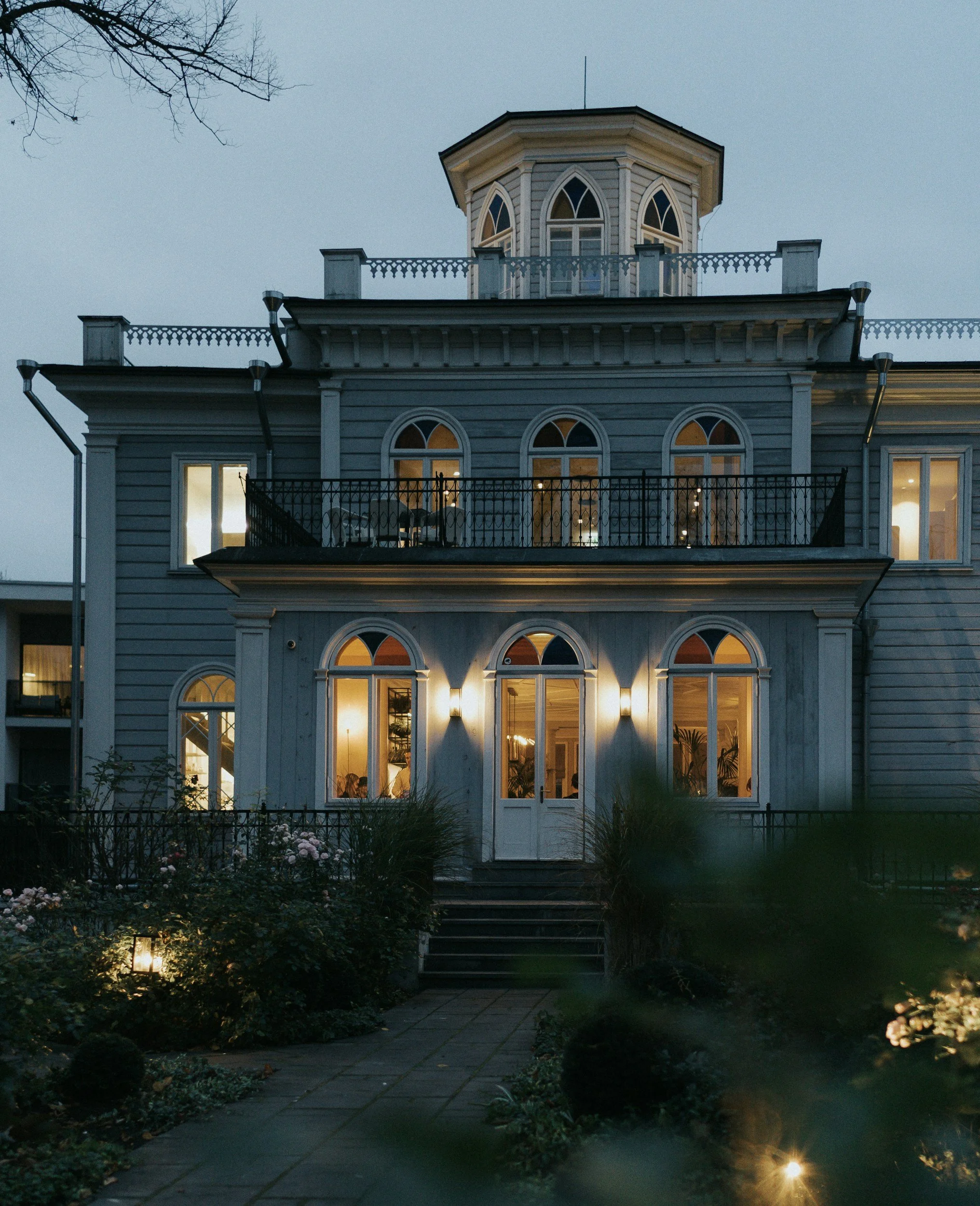 A multi-story house illuminated at dusk, with arched windows, a balcony, and a garden in the front.