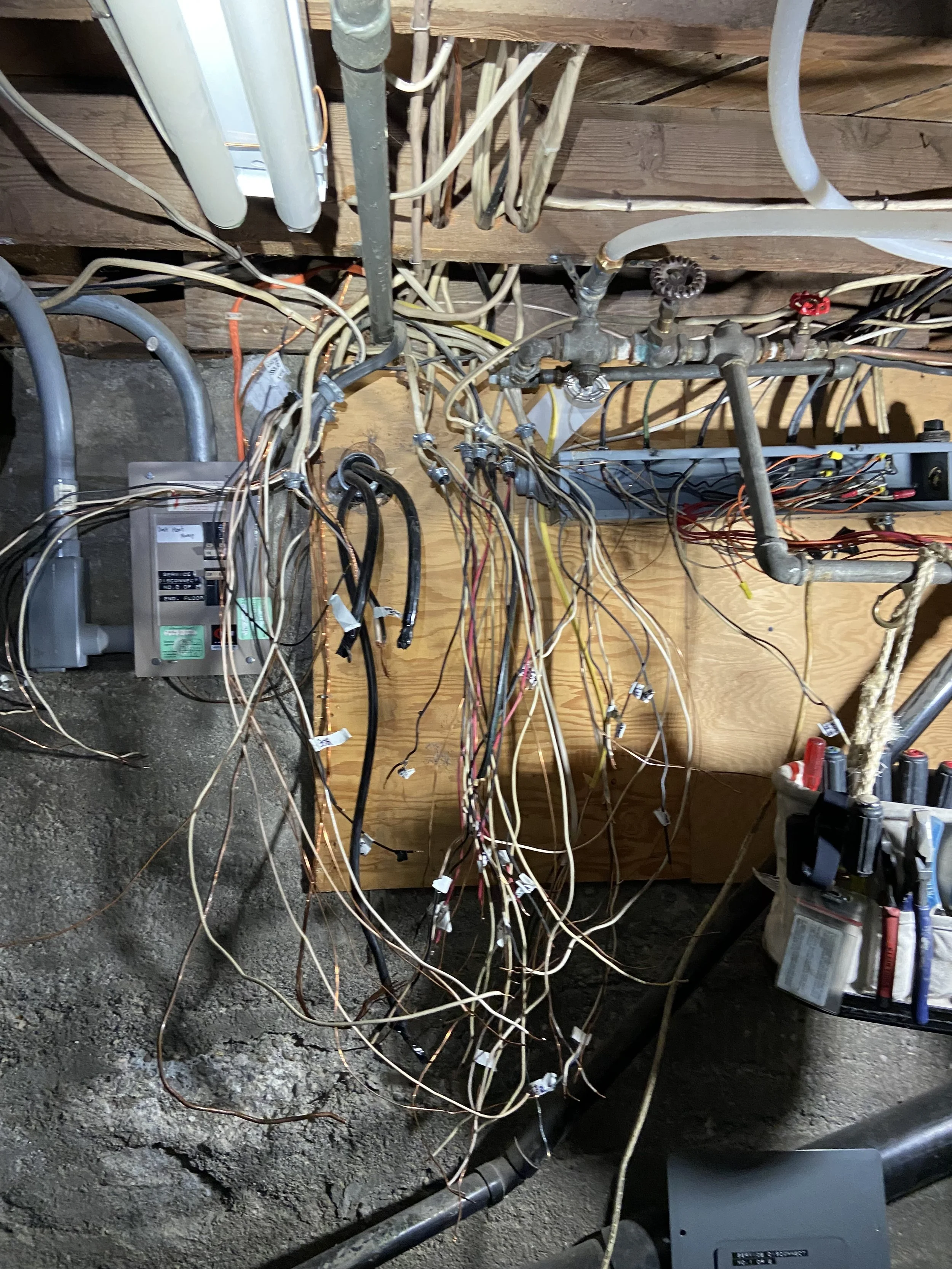 A messy collection of electrical wires and plumbing pipes in a basement or crawl space, with some connections wrapped in tape and others hanging loosely.