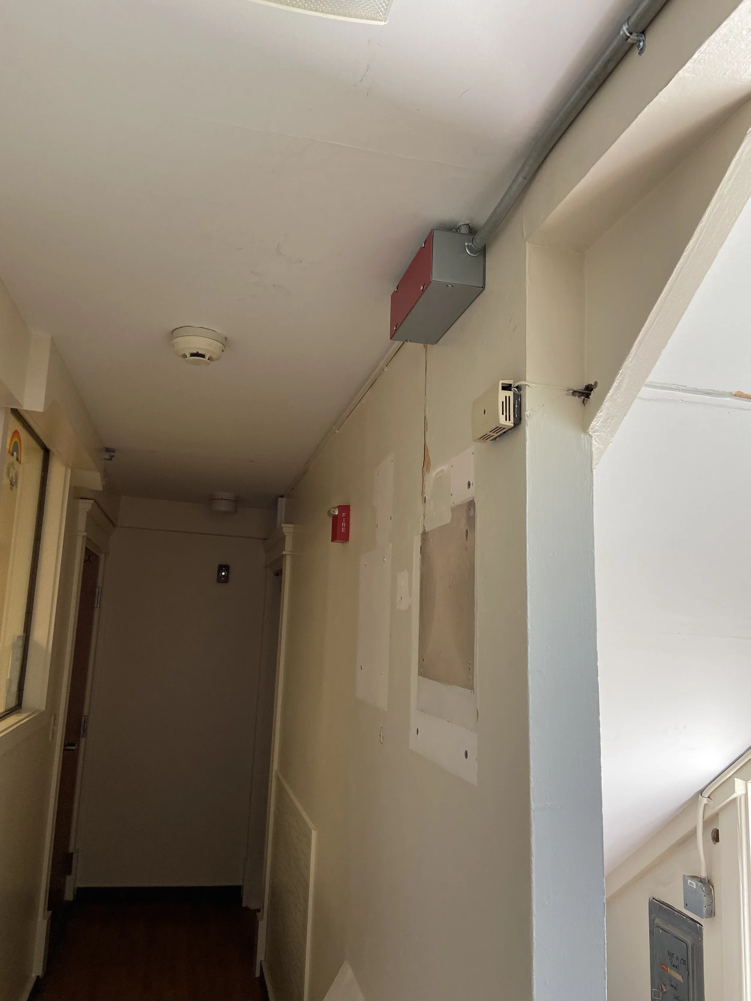 Empty hallway with exposed electrical wiring, conduit, and junction box on wall, smoke detector on ceiling, fire alarm on wall, and a partially covered wall panel.