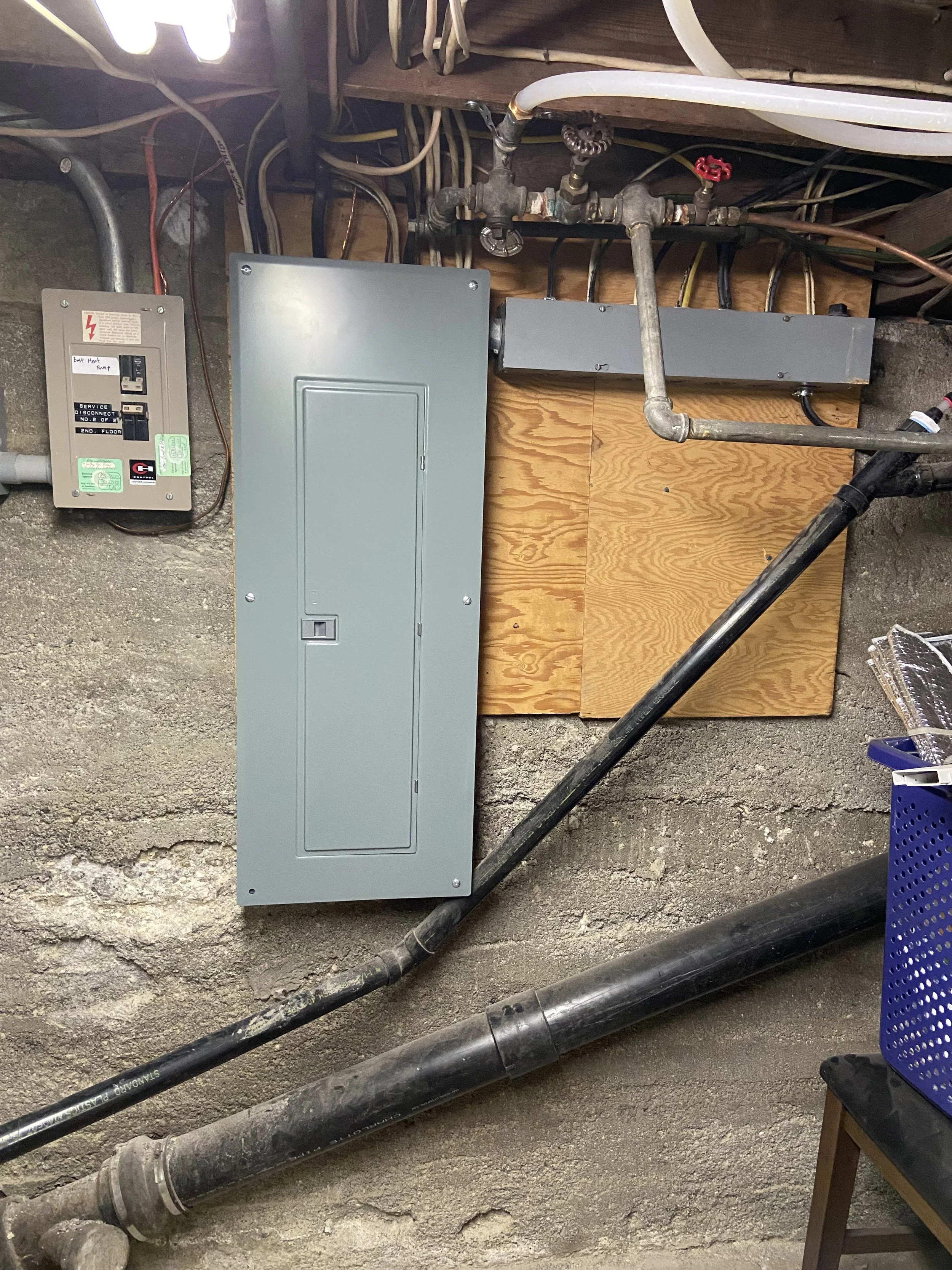 Electrical panel, plumbing pipes, and circuit breaker box on a concrete wall in a basement or utility room.