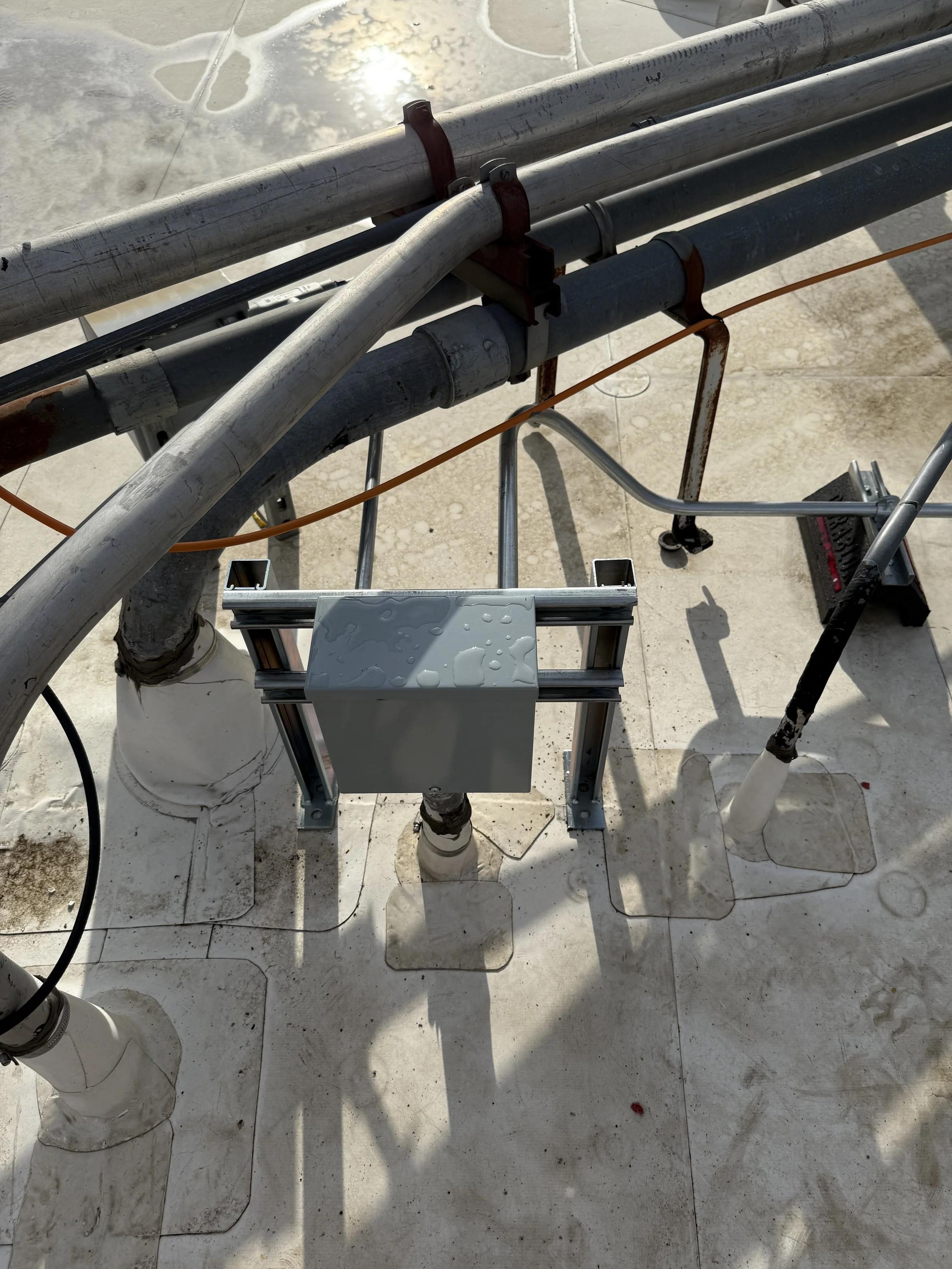 Multiple metal pipes, a small metal box with water droplets, and electrical wires on a concrete surface with puddles.