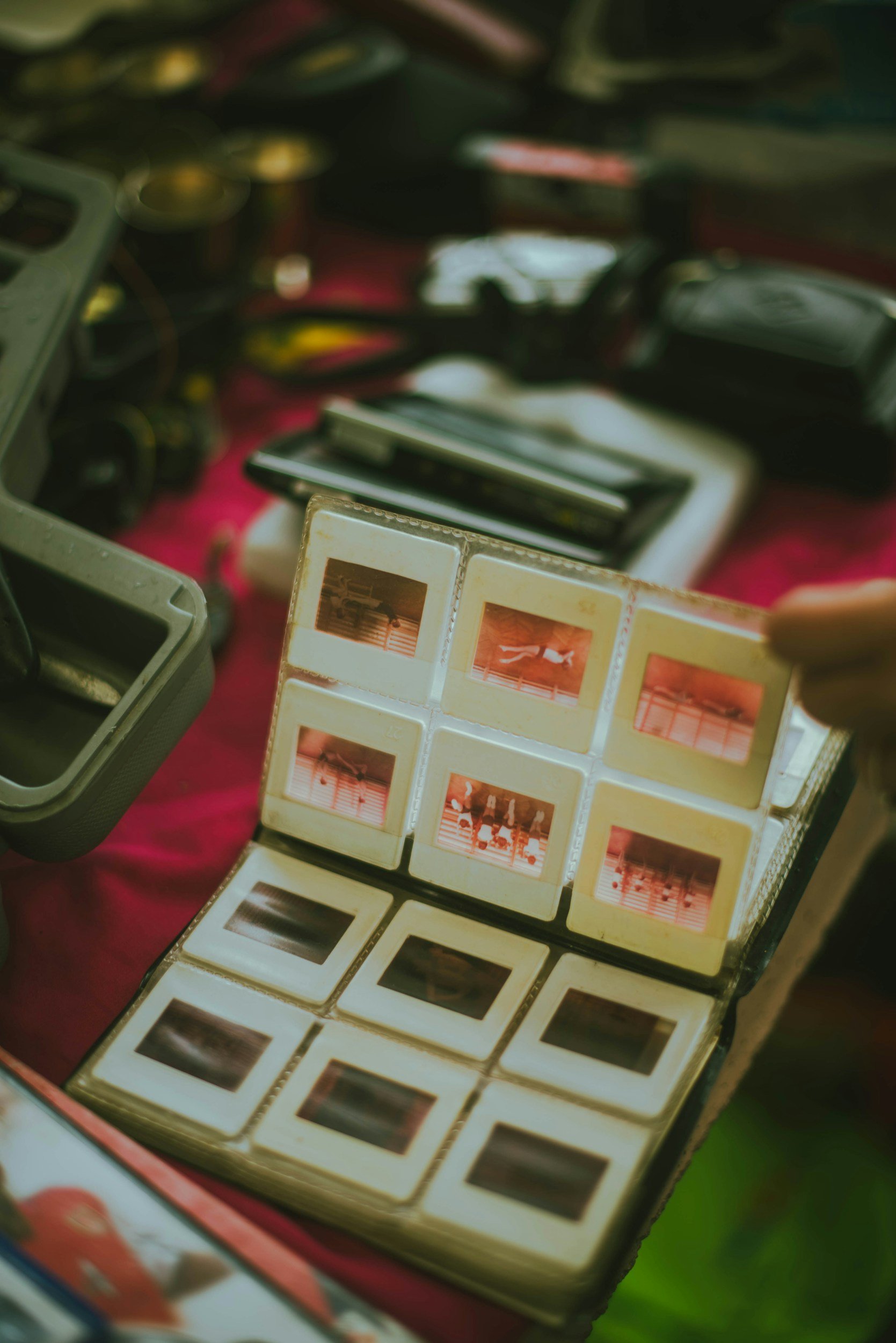 A vintage photo album with multiple small photographs, some showing people and activities like swimming, placed among other camera equipment on a red cloth surface.
