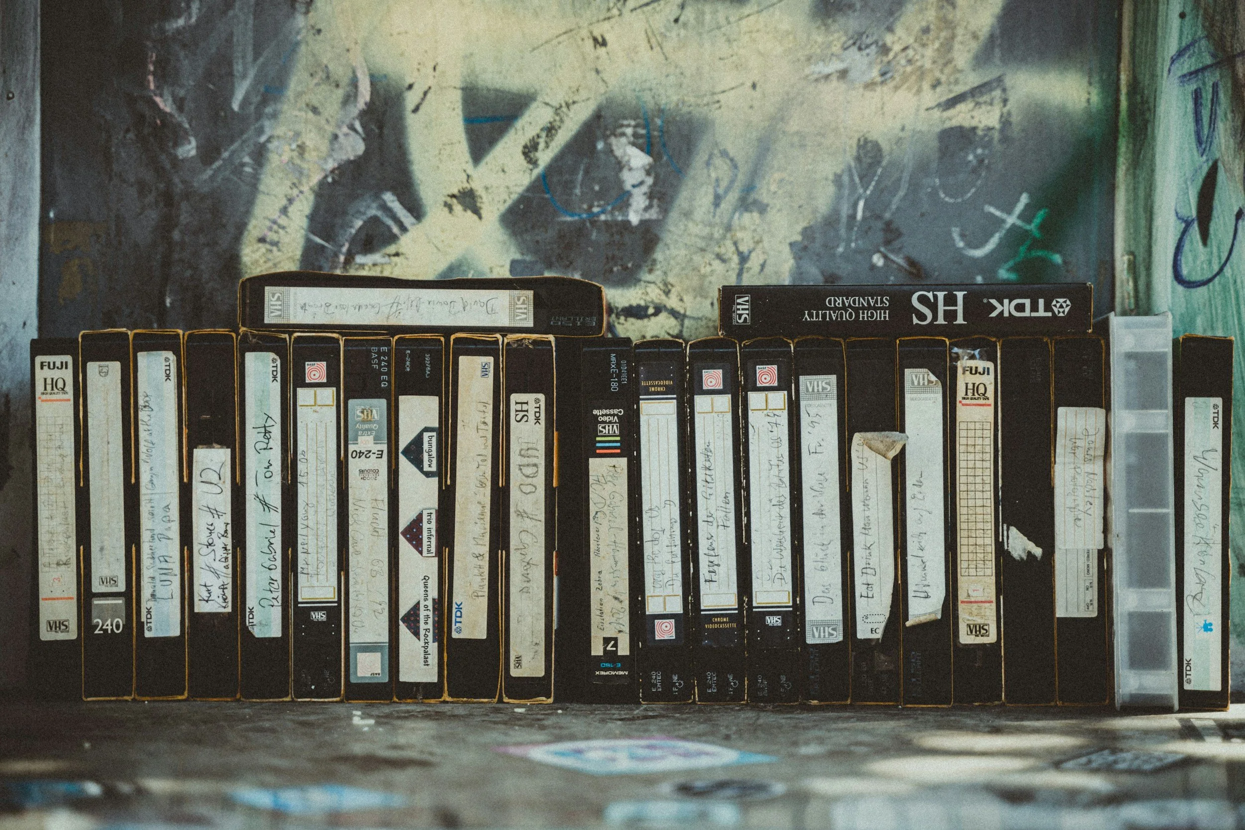 A collection of vintage VHS tapes arranged on a surface against a graffiti-covered wall.