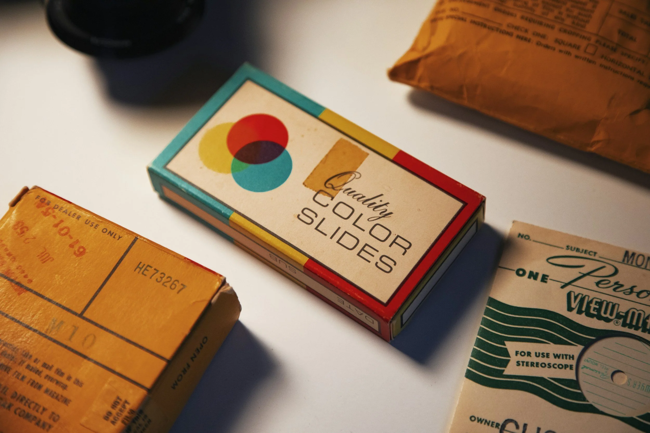 Three vintage film items on a white surface: a box of color slide film labeled 'Quadify Color Slides', a yellow box of film or photographic paper, and a green and white View-Master reel box for stereoscopes.