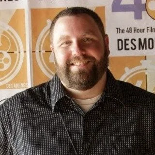 A bearded man smiling in front of a backdrop with logo and text, possibly at an event or interview.