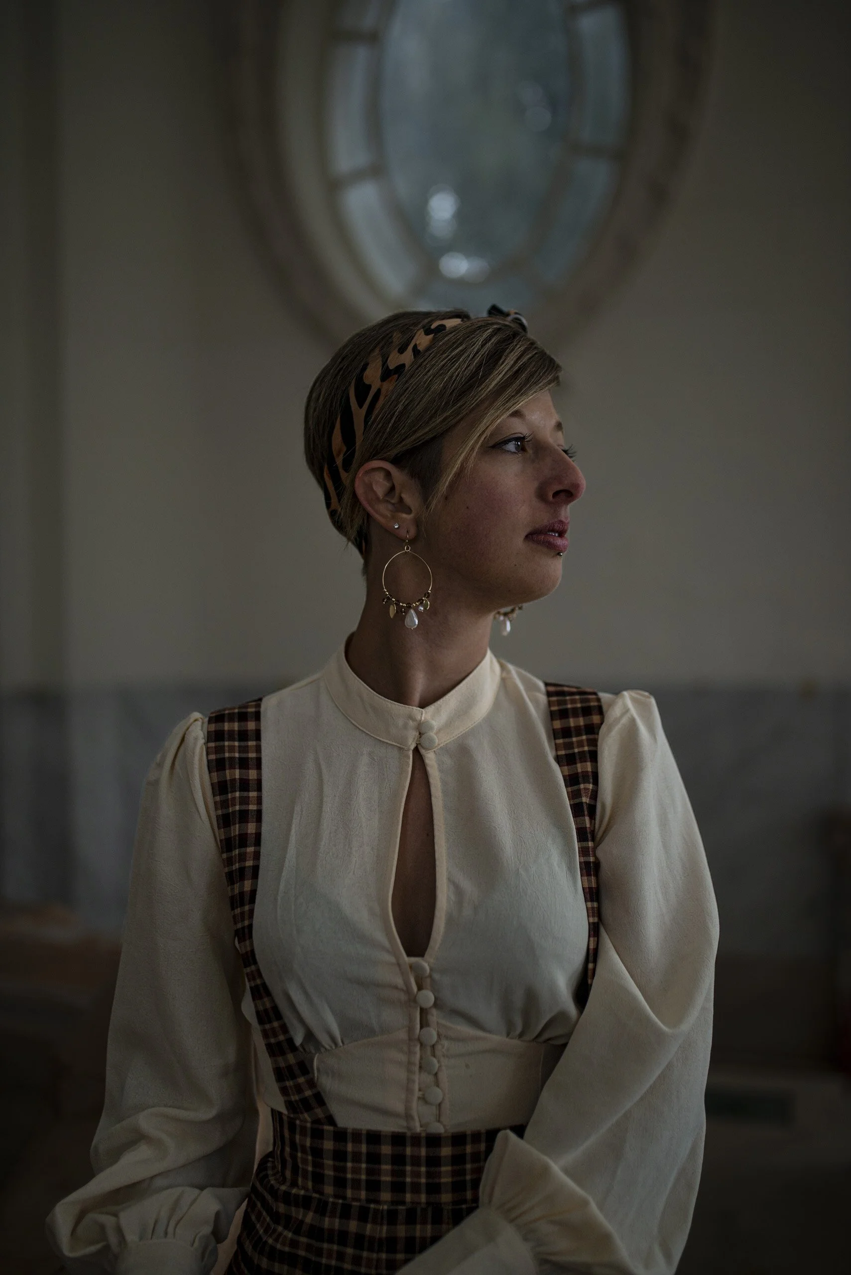 A woman with short hair, wearing a leopard print headband, a cream-colored blouse with a keyhole opening, and plaid suspenders, standing indoors with a round window in the background.