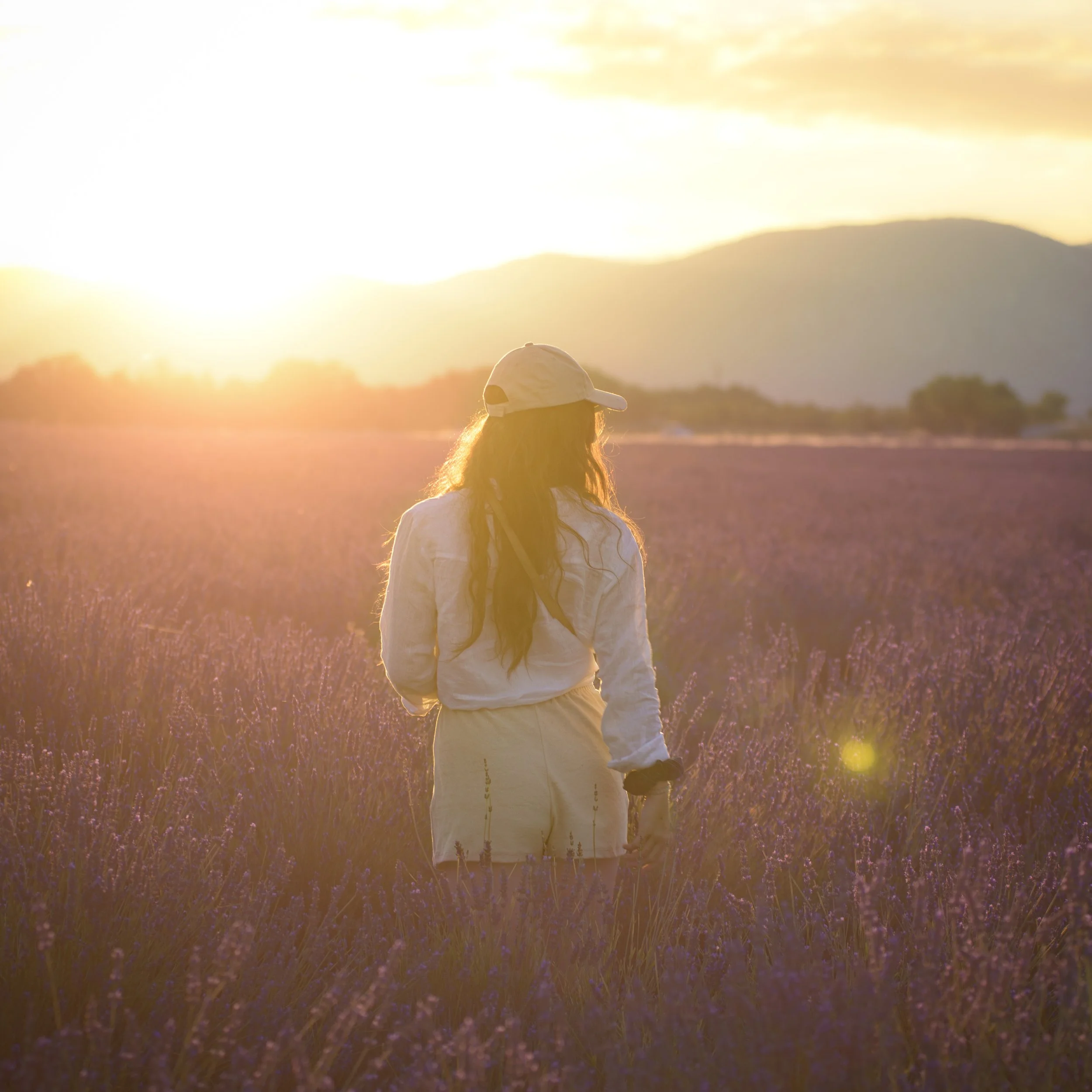 A woman standing in a lavender field during sunset, wearing a white cap and white outfit.