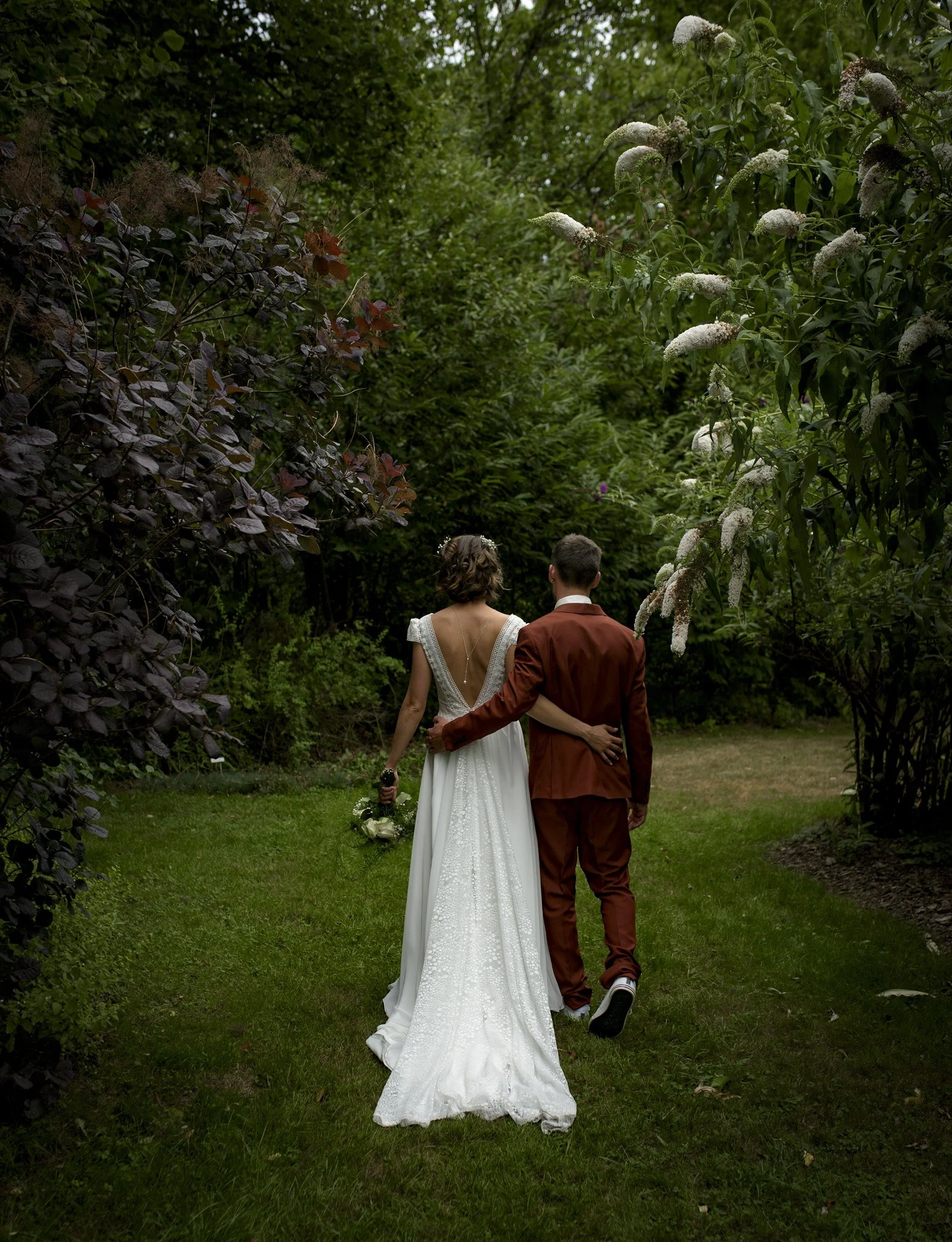 A couple in wedding attire walking through a lush garden, viewed from behind. The woman is in a white wedding dress holding a bouquet, and the man is in a brown suit with his arm around her waist.