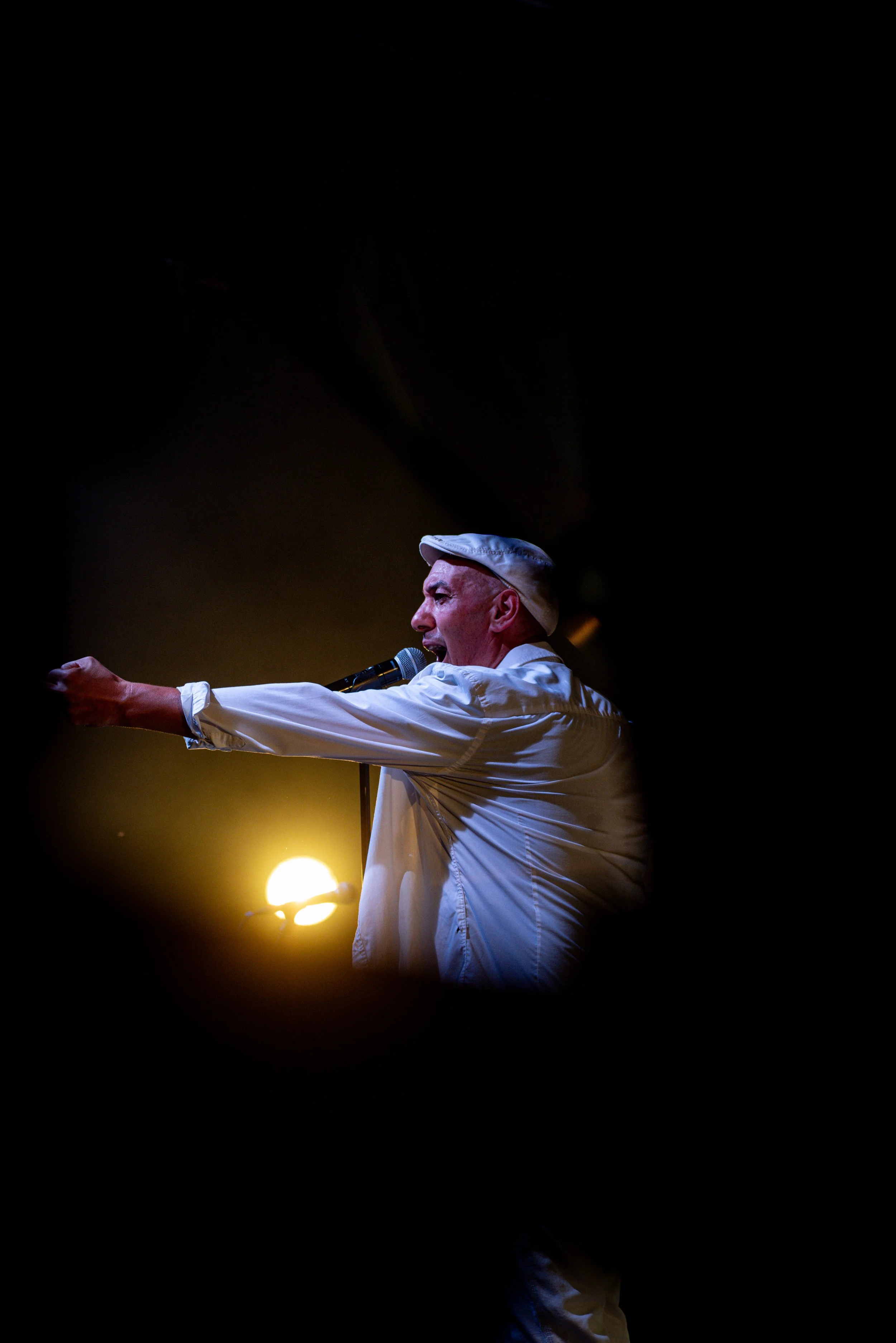 Performer singing into a microphone on stage, wearing a white shirt and hat, with a bright light behind him.