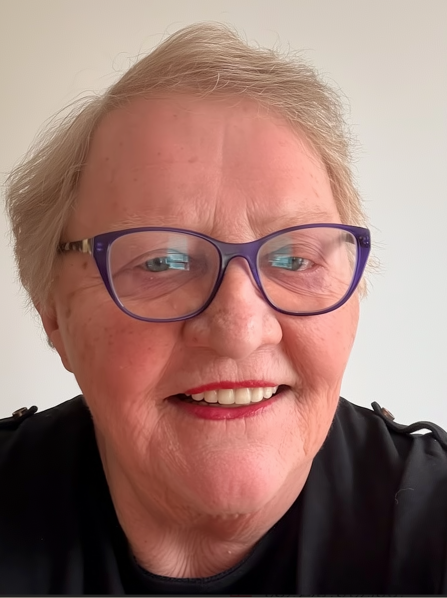 A close-up selfie of a smiling older woman wearing purple glasses and a black shirt, standing against a plain light-colored background.
