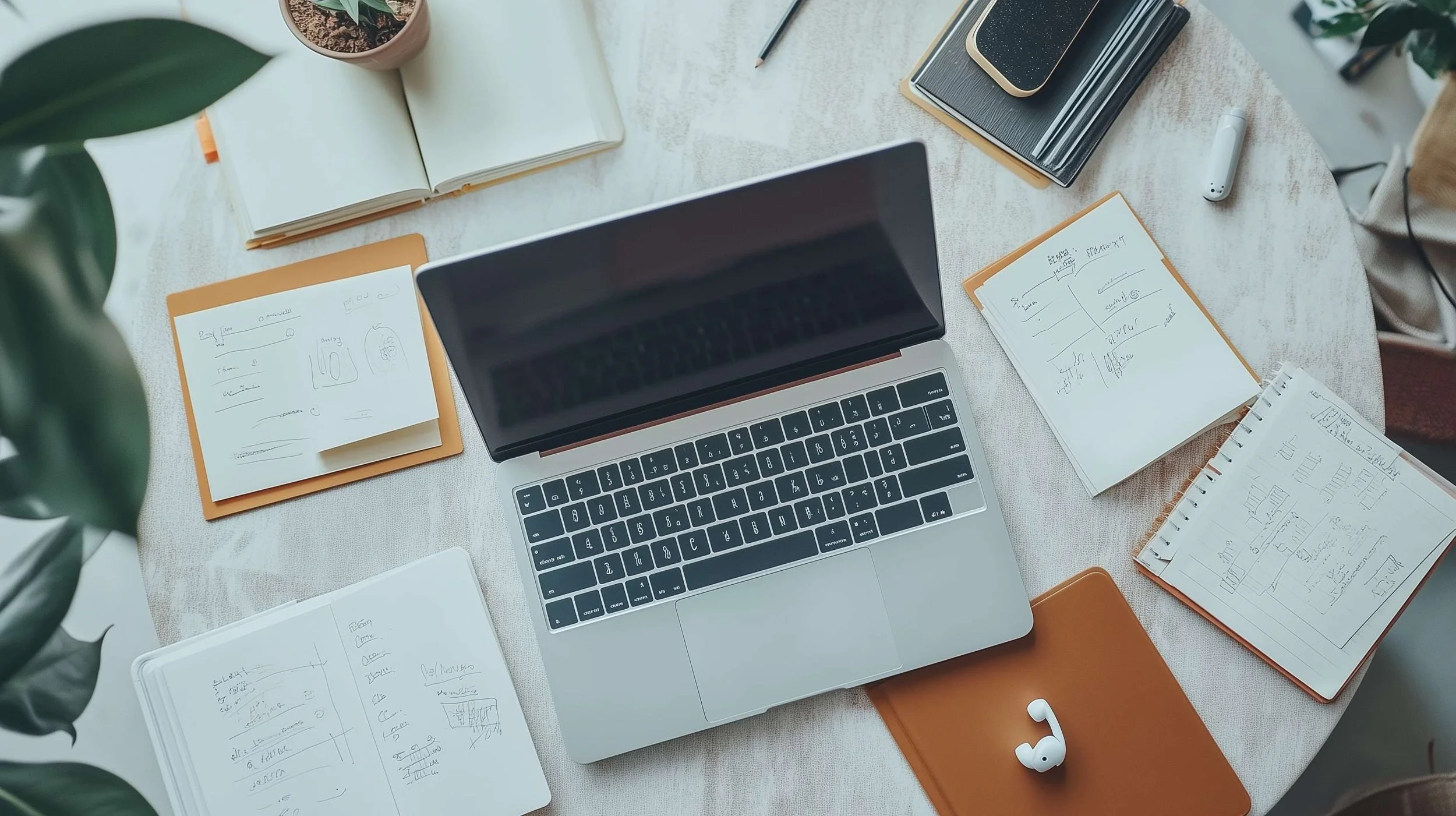 A table top with an open laptop, earbuds and notes, notebooks scattered around.