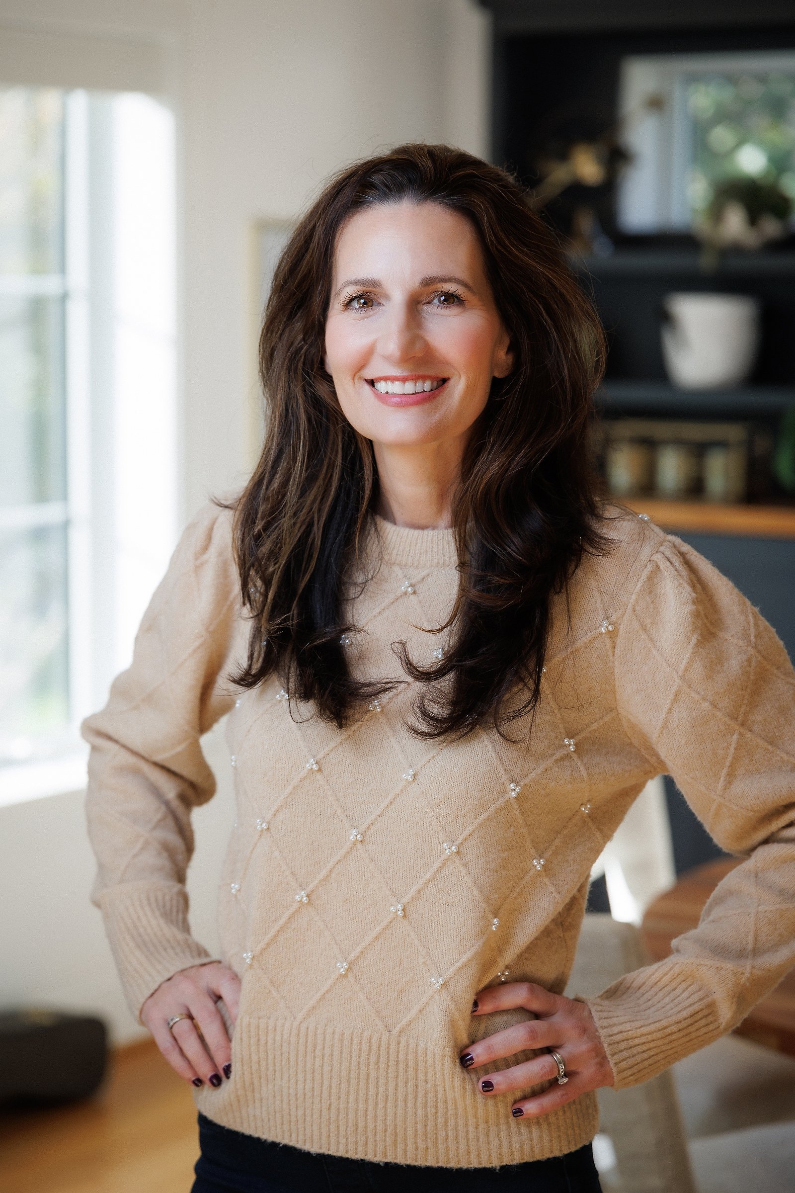 A smiling woman with long, wavy dark hair, wearing a beige sweater with pearl embellishments, standing in a well-lit room with a window and shelf in the background.