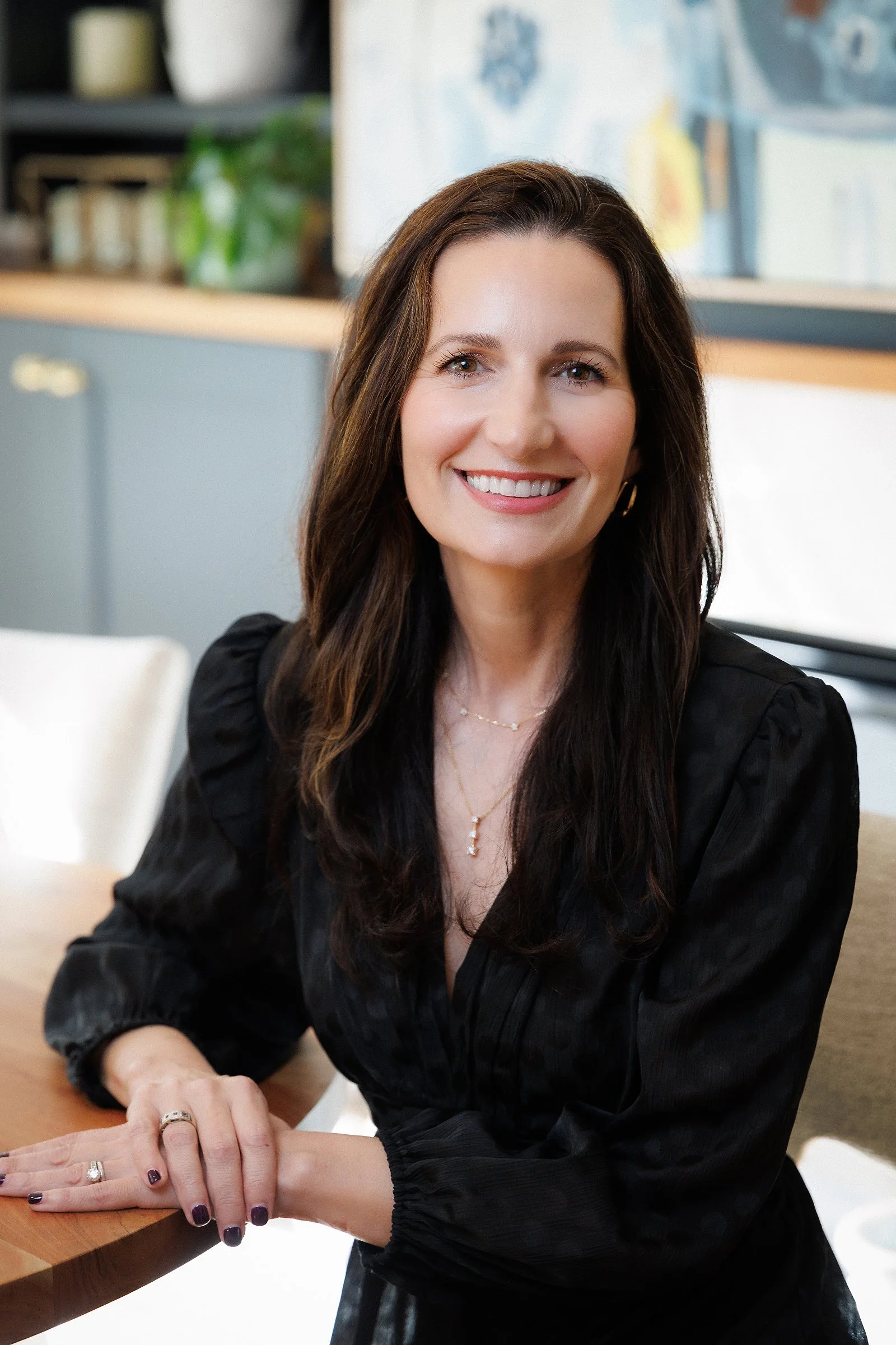 A smiling woman with long brown hair, wearing a black blouse and jewelry, sitting at a wooden table in a modern kitchen.