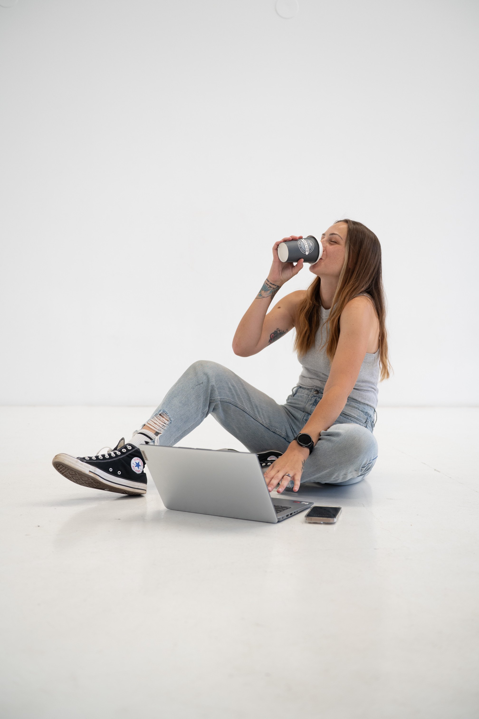 A woman sitting on the floor, drinking from a cup, with a laptop and smartphone nearby, in a minimal studio setting.