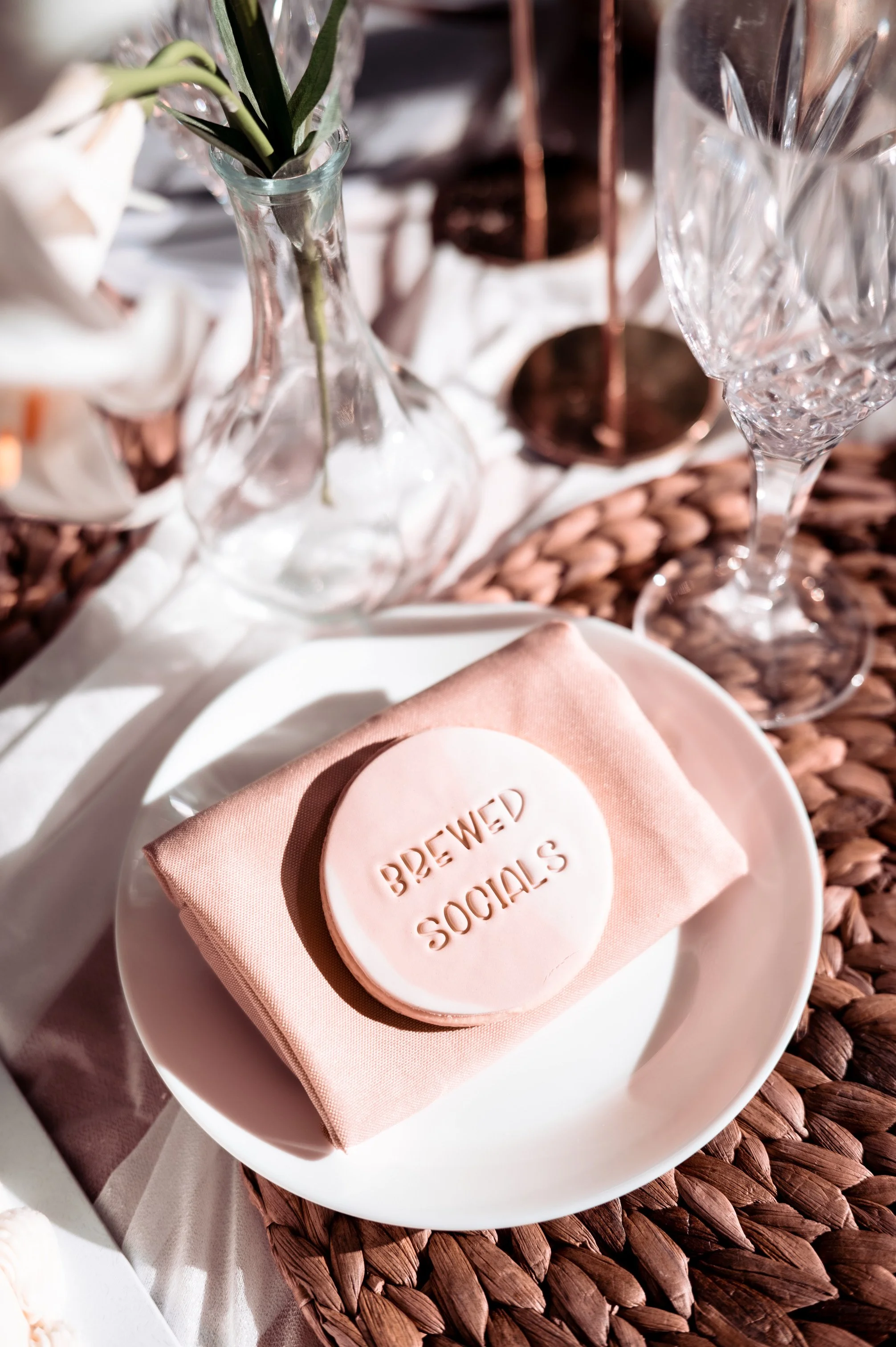 Table setting with a pink napkin, a pink soap with the words "BREWED SOCIALS" embossed, with glassware and a decorative glass vase with plant stems in the background. From a free female small business owners event.