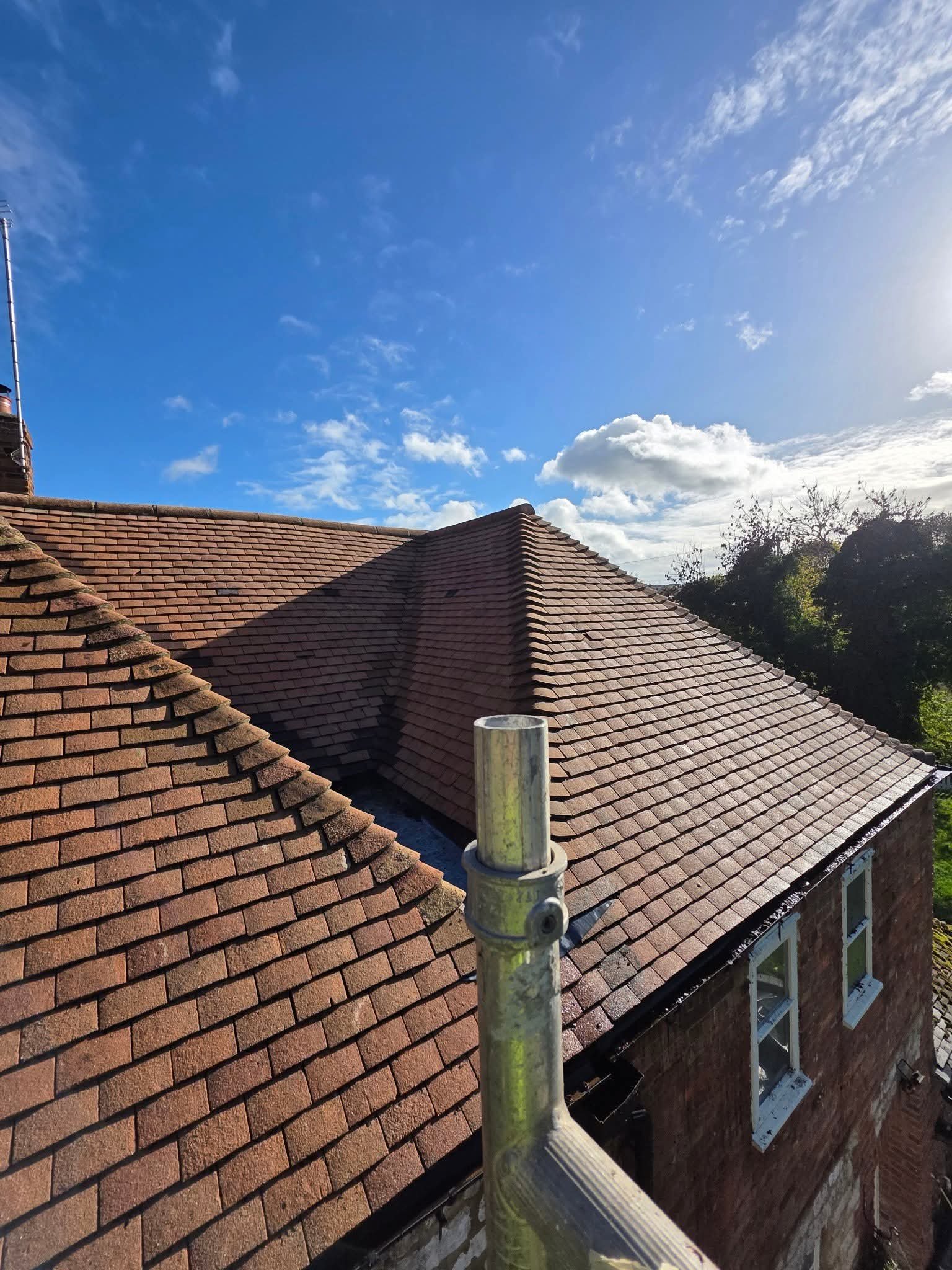 Residential roof with reddish-brown shingles, clear blue sky with scattered clouds, part of a chimney on the left, a chimney pipe and scaffolding in the foreground, trees in the background.