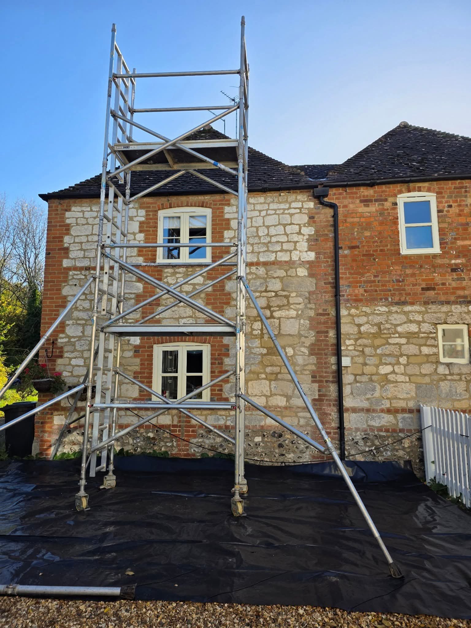 Scaffolding in front of a brick house with two white-framed windows, a roof with dark shingles, and a black drainage pipe. The ground is covered with black tarp. The sky is clear and blue.