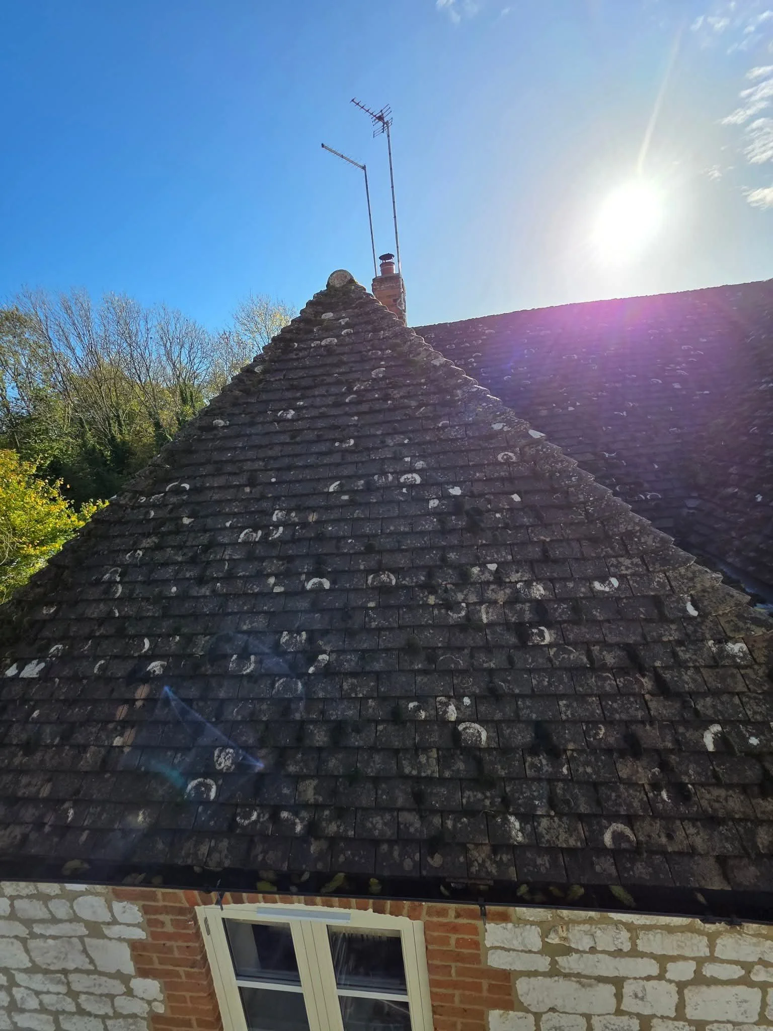 A photograph taken during daytime shows the roof of a brick house with weathered, moss-covered shingles. A chimney with an antenna and television aerials extends from the roof against a bright blue sky with the sun shining brightly. Trees with sparse foliage are visible in the background.