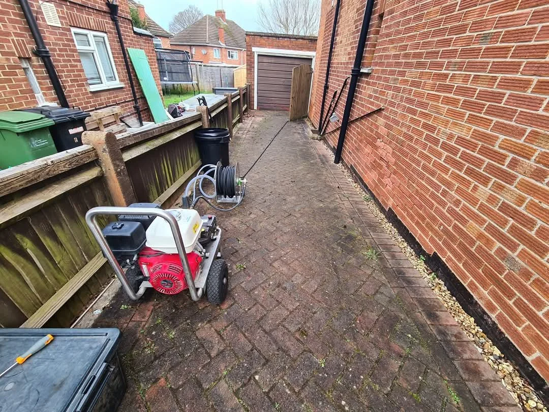 Dirty brick driveway covered in dark moss and mud with a professional jet washer machine on-site in Woodley before cleaning.