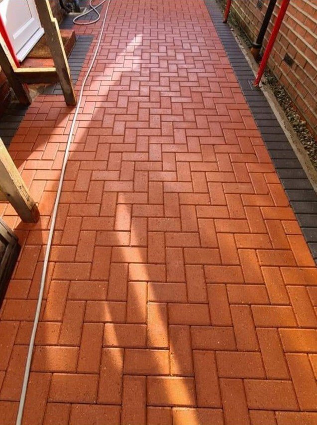 Red brick walkway with shadow and black border around the edge. Part of a wooden structure and a garden hose are visible on the left side. A brick wall with red support beams is on the right side.