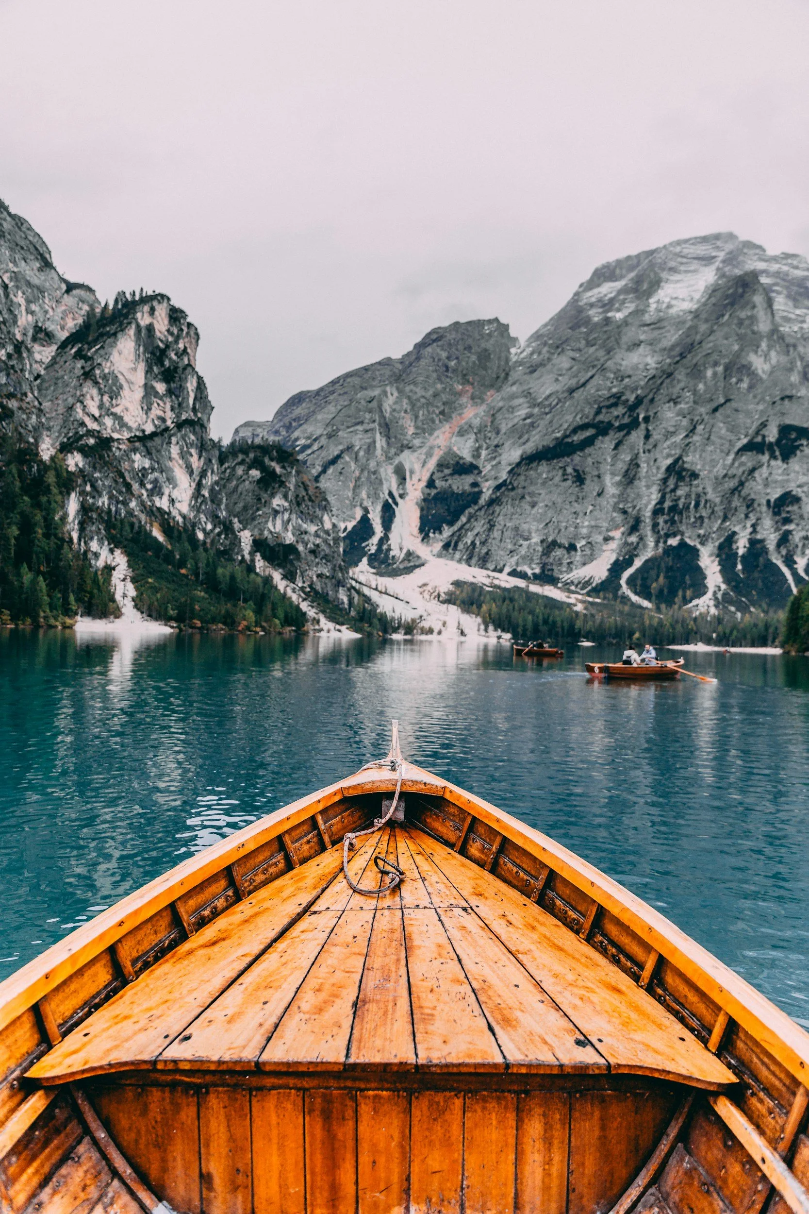 View from the front of a wooden boat on a calm lake surrounded by tall mountains and trees, with other boats floating nearby.