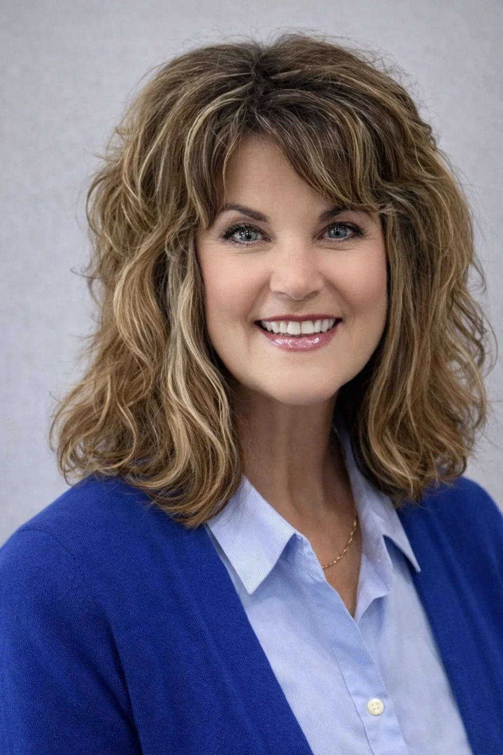 A smiling woman with curly, shoulder-length light brown hair, wearing a blue blazer over a light blue button-up shirt, looking directly at the camera.