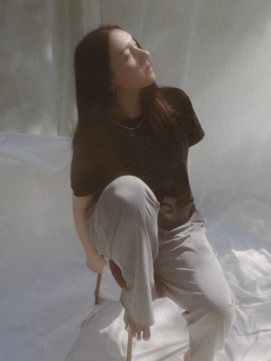 A person with long hair sitting on a chair with one leg raised, looking upwards with closed eyes against a plain background.