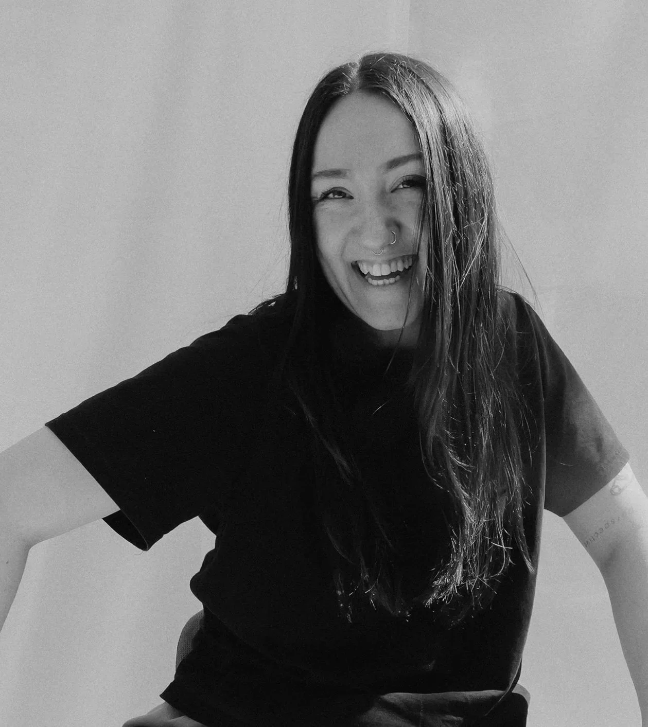 Black and white photo of a woman with long dark hair, smiling and showing teeth, wearing a black t-shirt, standing in front of a plain wall.