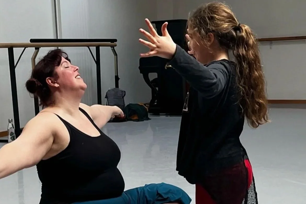 A woman in black top and blue pants smiling with arms outstretched, facing a girl with curly hair in dark clothing, who is raising her hands and appears to be playing or interacting with her.
