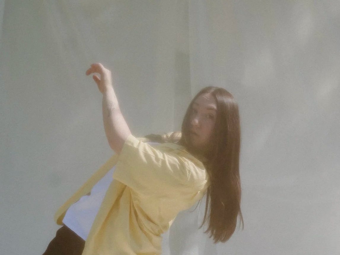 A young woman with long brown hair taking a selfie, wearing a yellow jacket over a white top, standing against a plain, light-colored wall.