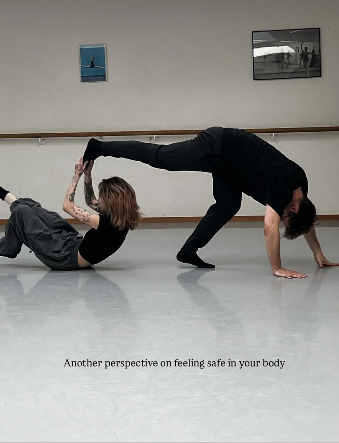 A man and woman are performing a complex acro yoga pose in a dance studio, with the man in a downward dog position and the woman holding his feet while seated on the floor.