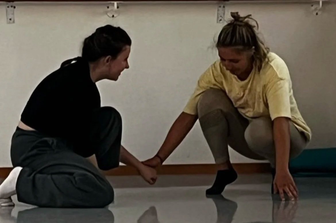 Two women are practicing acro yoga indoors on a shiny floor. One woman is kneeling and holding the foot of the other woman, who is balancing on her hands and feet with one hand on the floor.