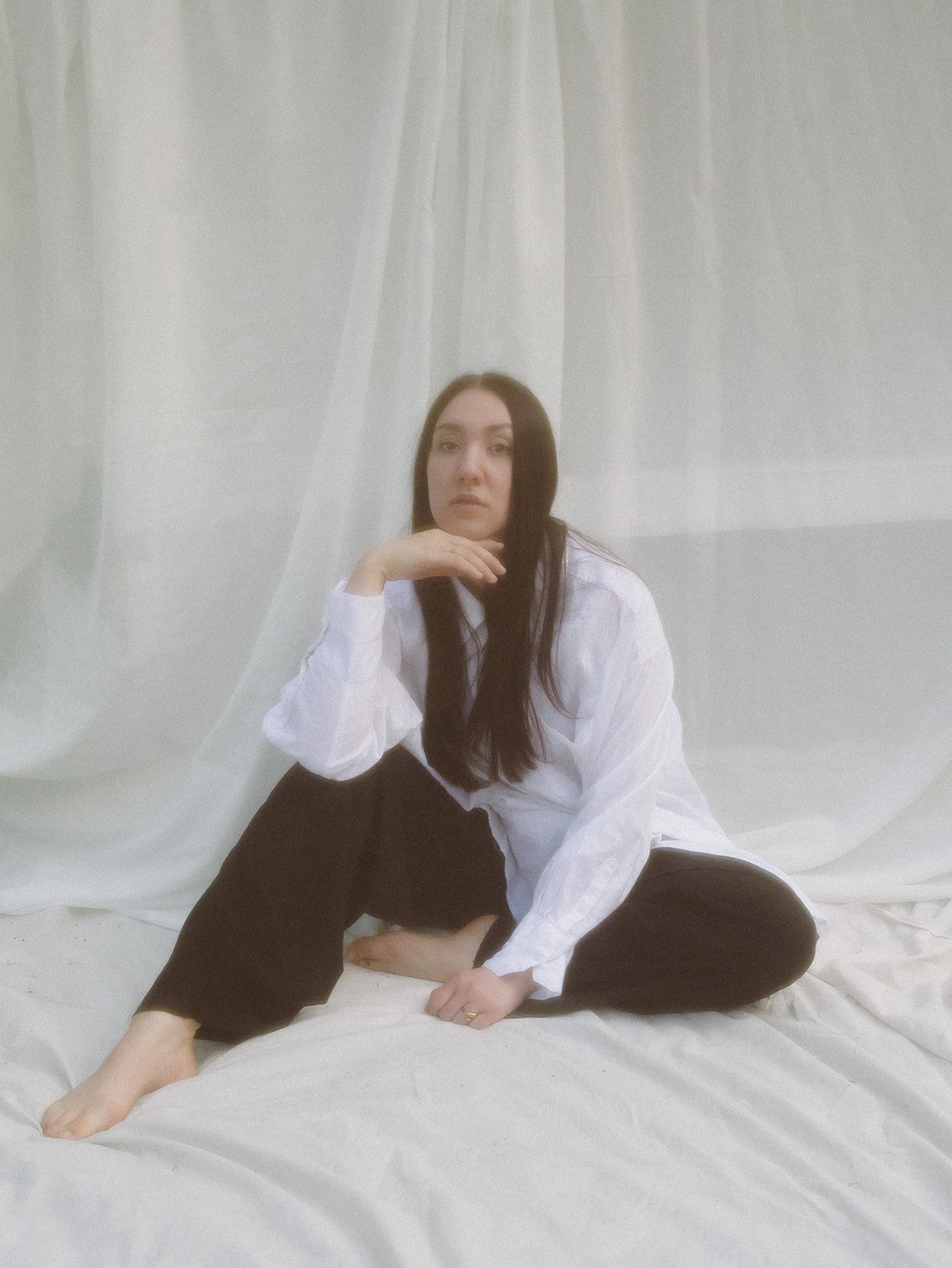 A woman with long dark hair sitting on the white fabric floor, dressed in a loose white shirt and black pants, with a white curtain backdrop.