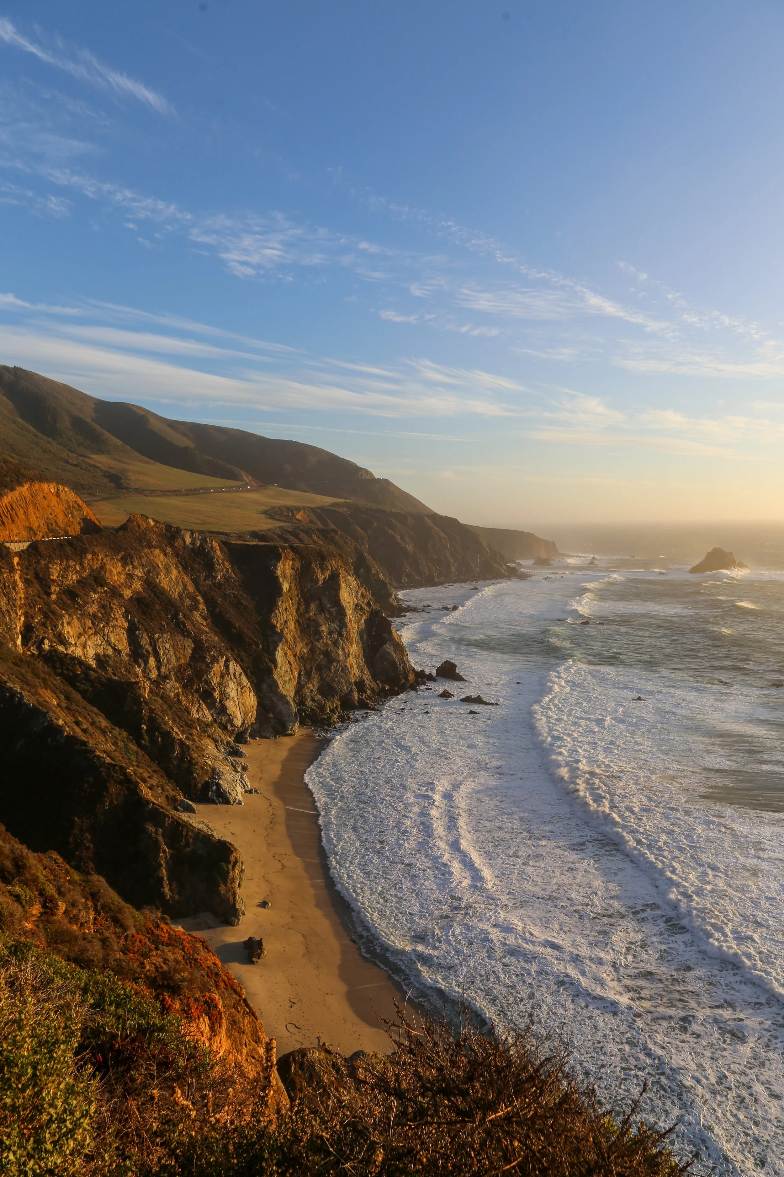 Sunset over rugged coastline with cliffs and waves crashing onto a small sandy beach.