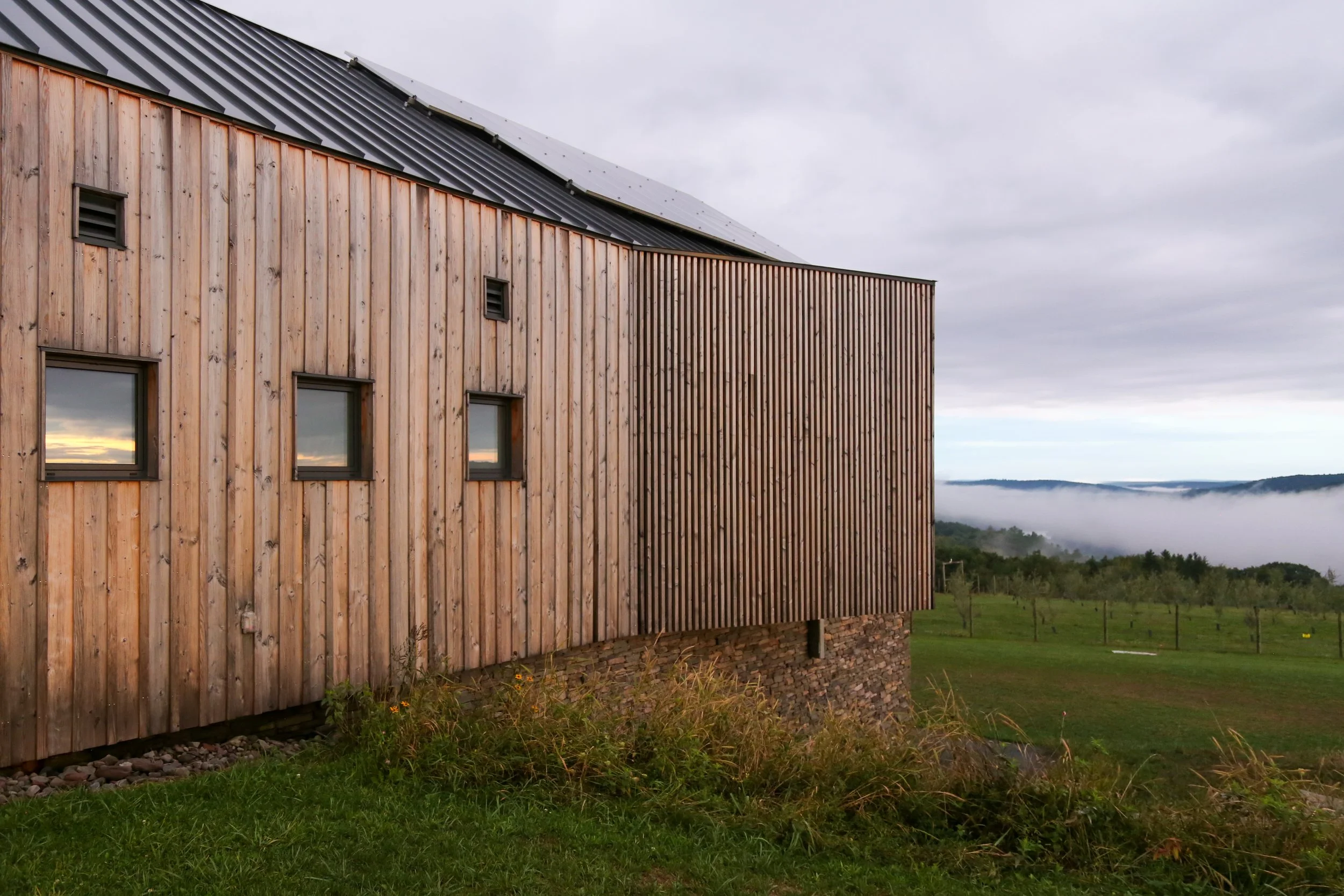 Modern house with wooden siding and small windows, situated on a grassy landscape with distant mountains and fog.