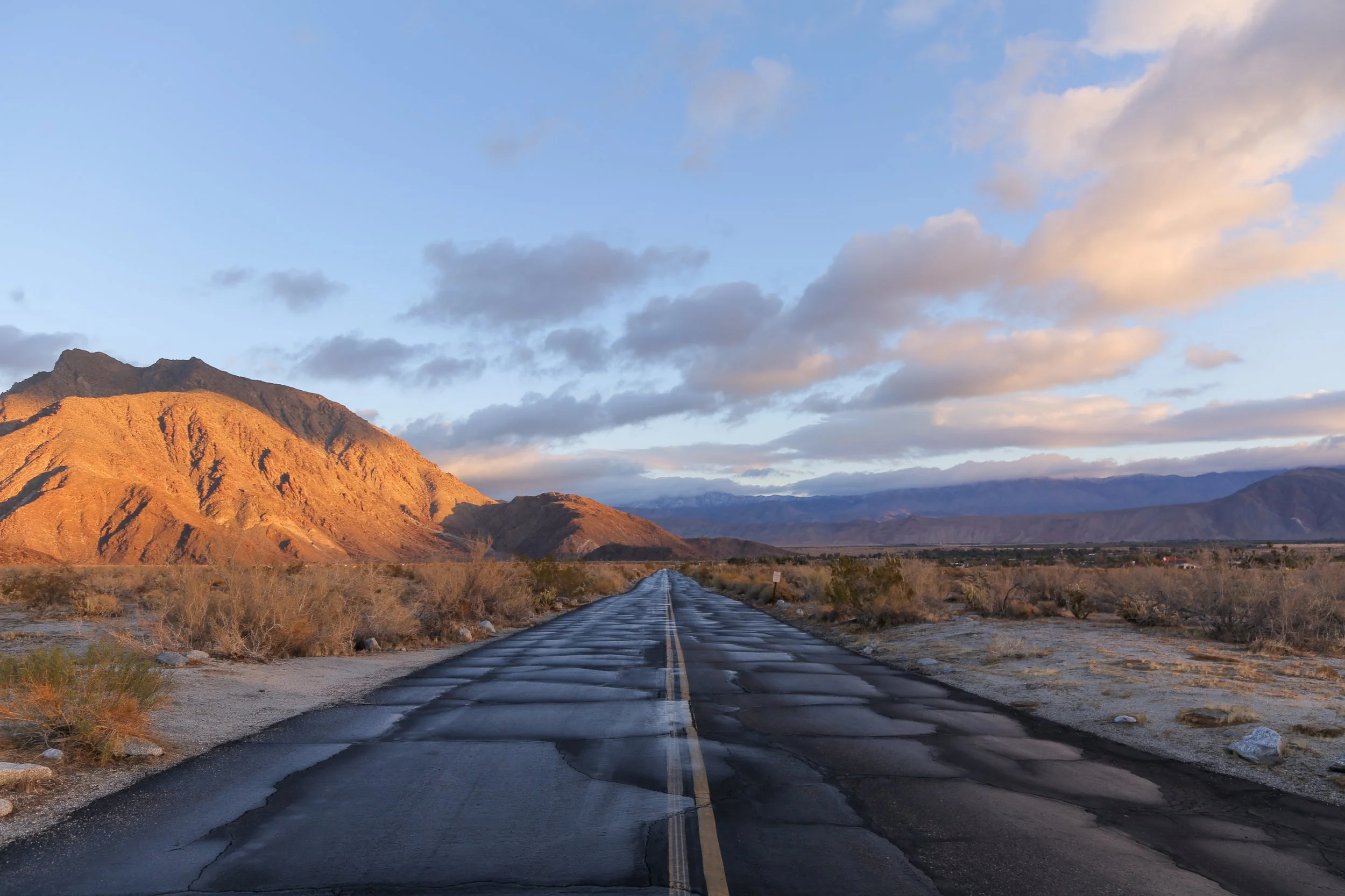 A deserted road stretches toward distant mountains at sunset, with dry desert vegetation on either side and a sky filled with scattered clouds.