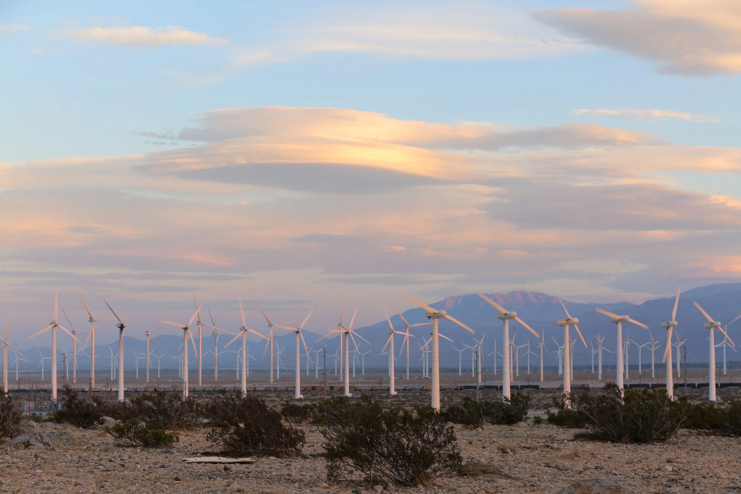 A landscape of numerous wind turbines in a desert with mountains in the background during sunset.