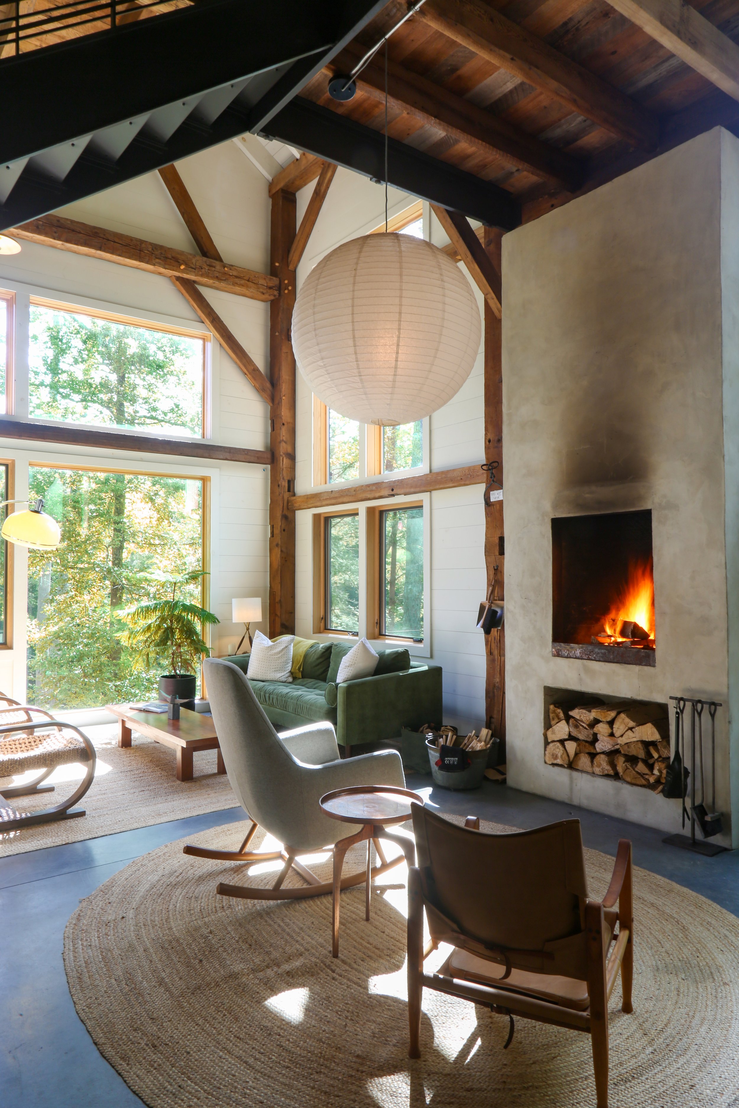 Living room with large windows, high ceiling with wooden beams, a fireplace with fire, a green sofa, a modern armchair, a lamp, a rug, and firewood stored below the fireplace, with natural light streaming in.