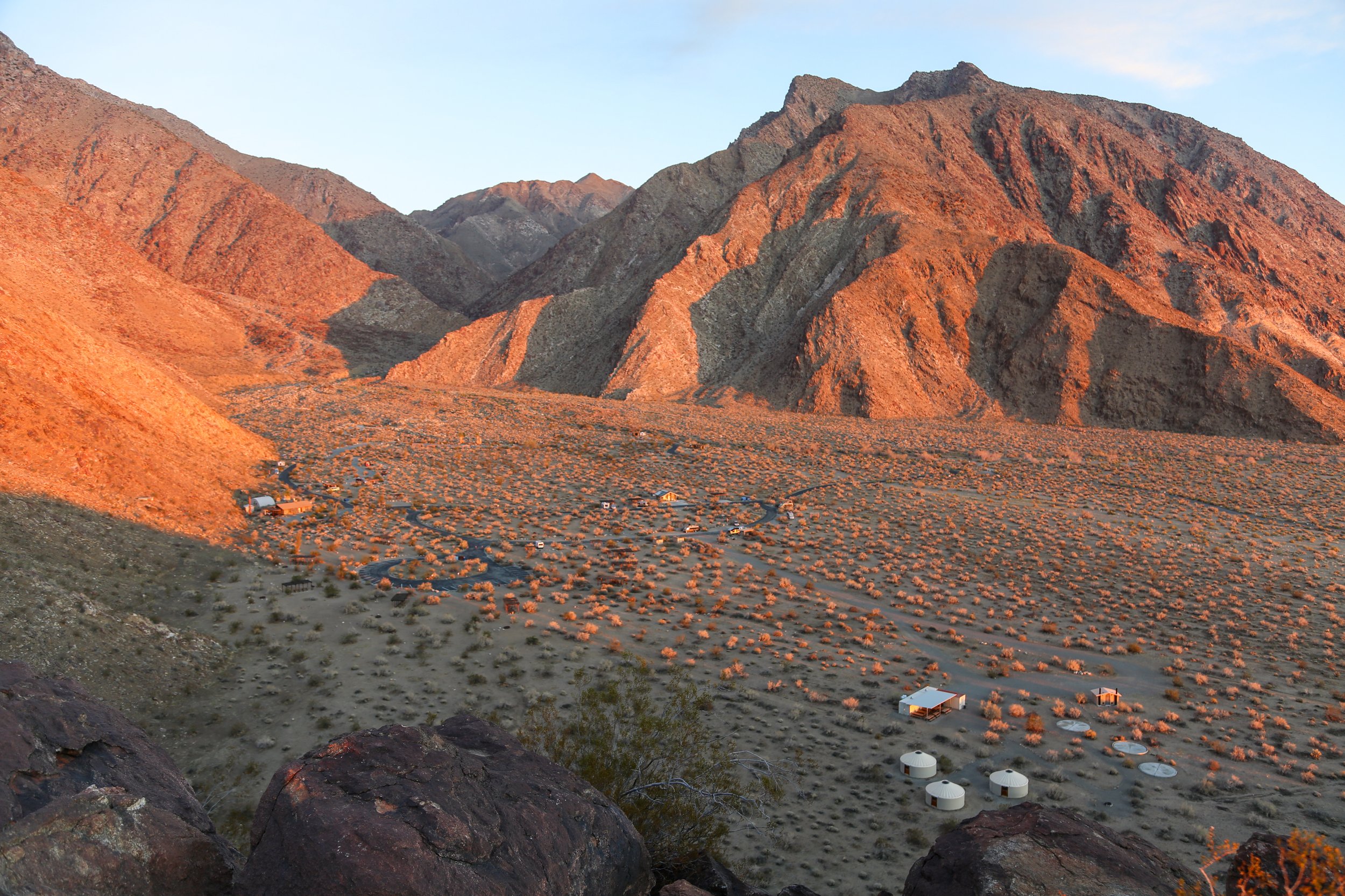 Desert valley with small buildings surrounded by reddish mountains at sunset.