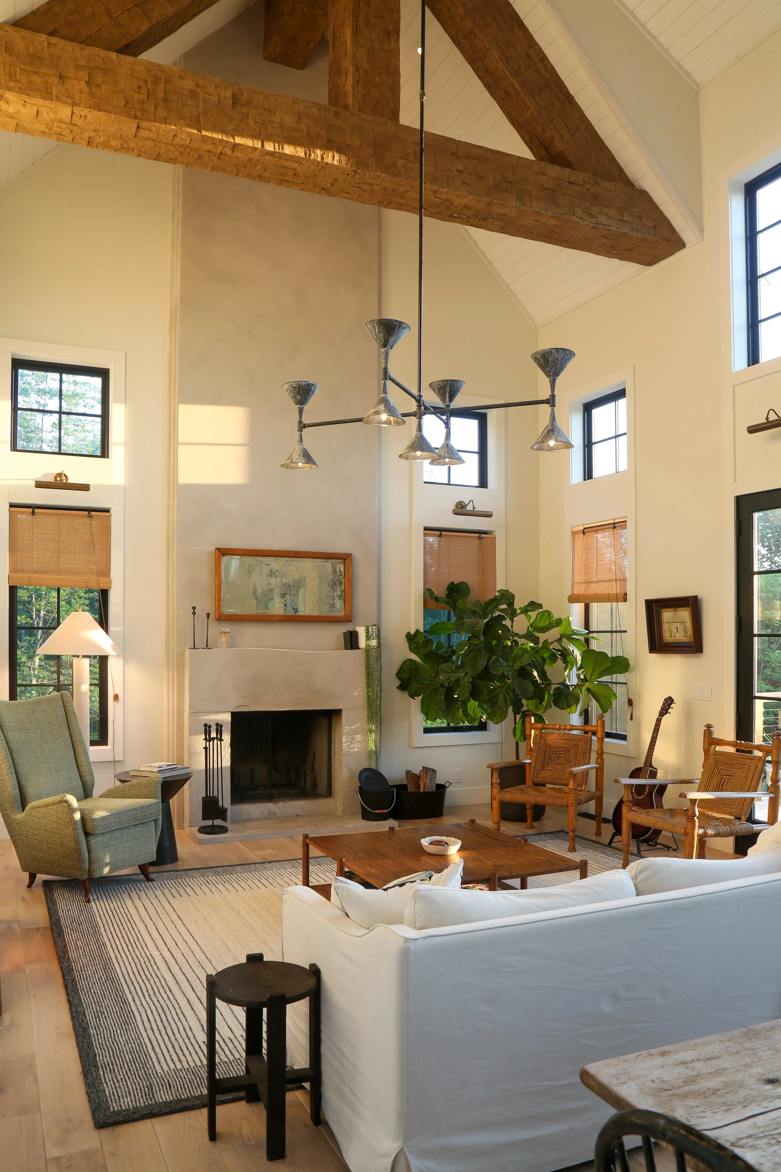 Living room with high ceilings featuring exposed wooden beams, large windows, a white sofa, a green armchair, a fireplace, a wooden coffee table, a black side table, a guitar, and houseplants.