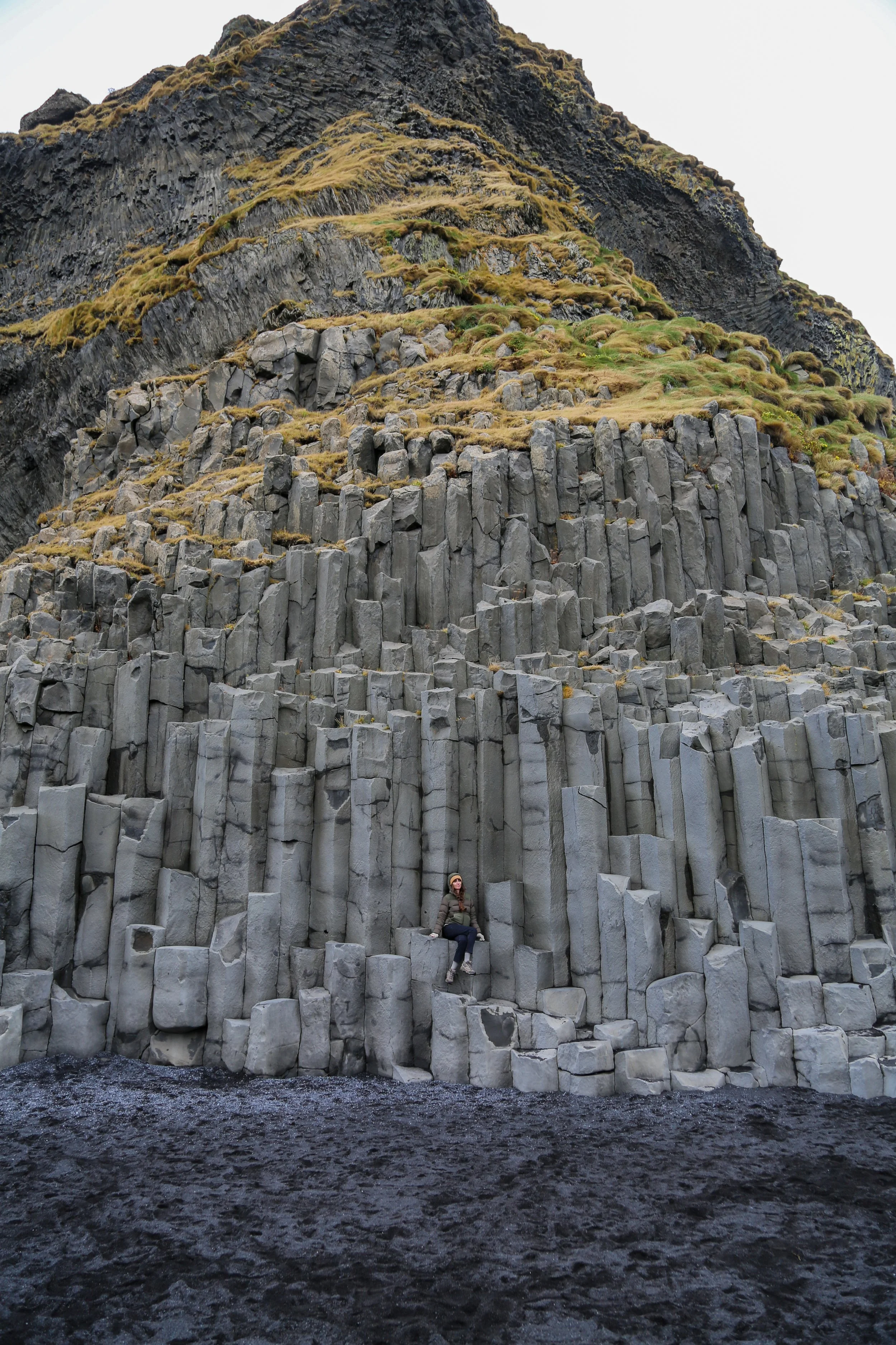 A woman sitting on basalt hexagonal columnar joints at a volcanic beach, with a mountain in the background.