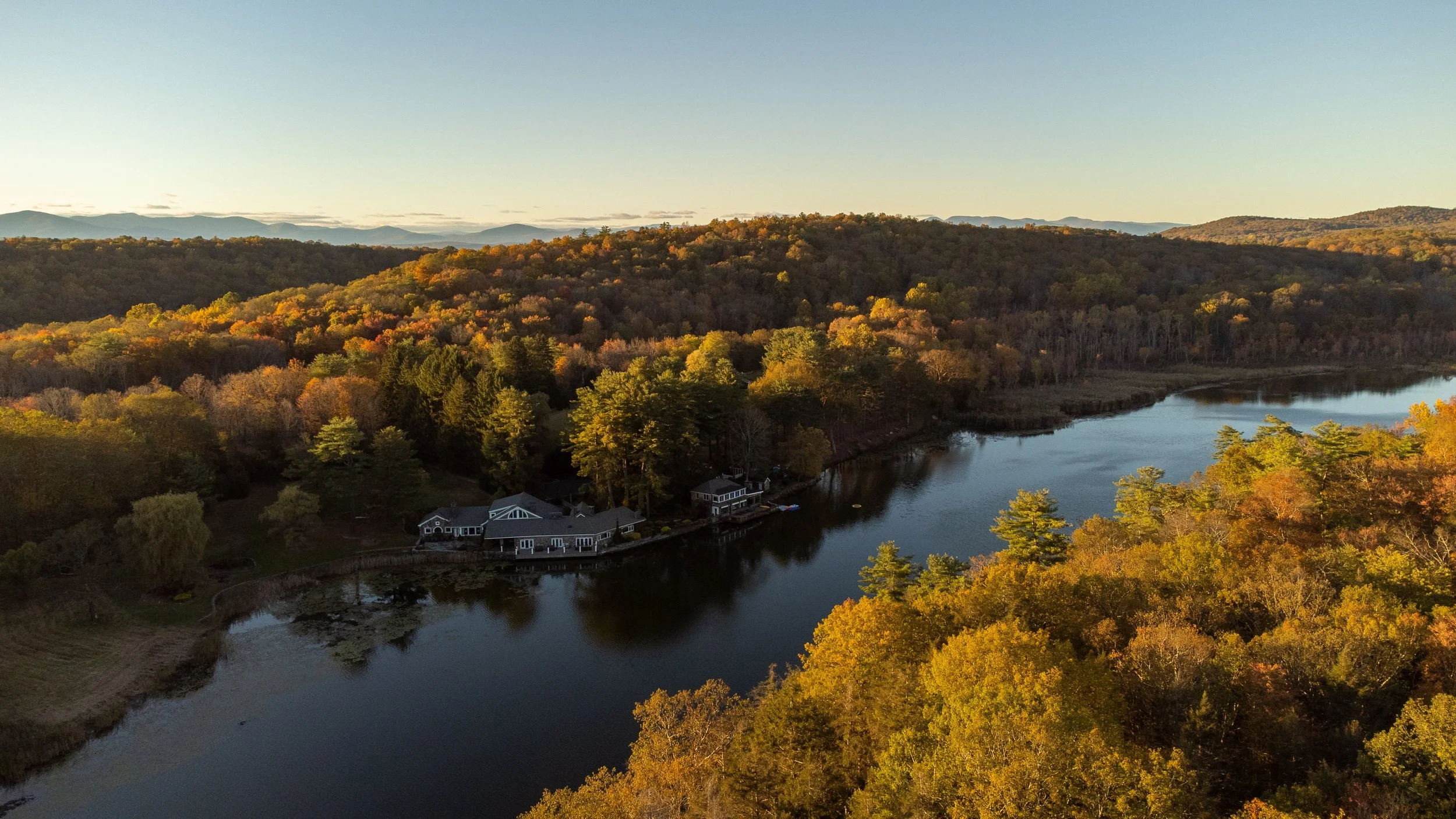 An aerial view of a lake surrounded by colorful autumn trees with a house on the shoreline, hills in the background, and a clear sky.