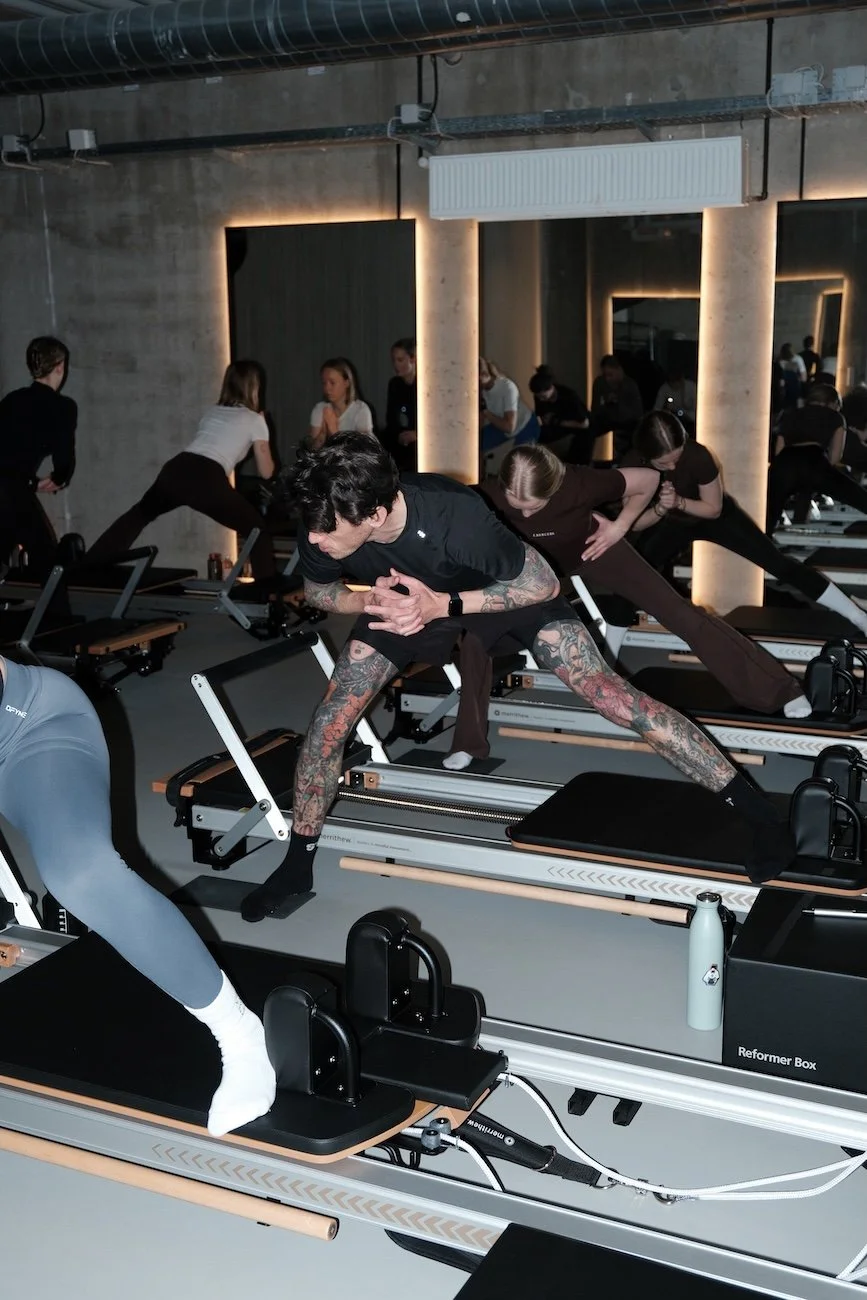 People participating in a Pilates class on reformer machines in a modern studio with concrete walls and mirror lighting.